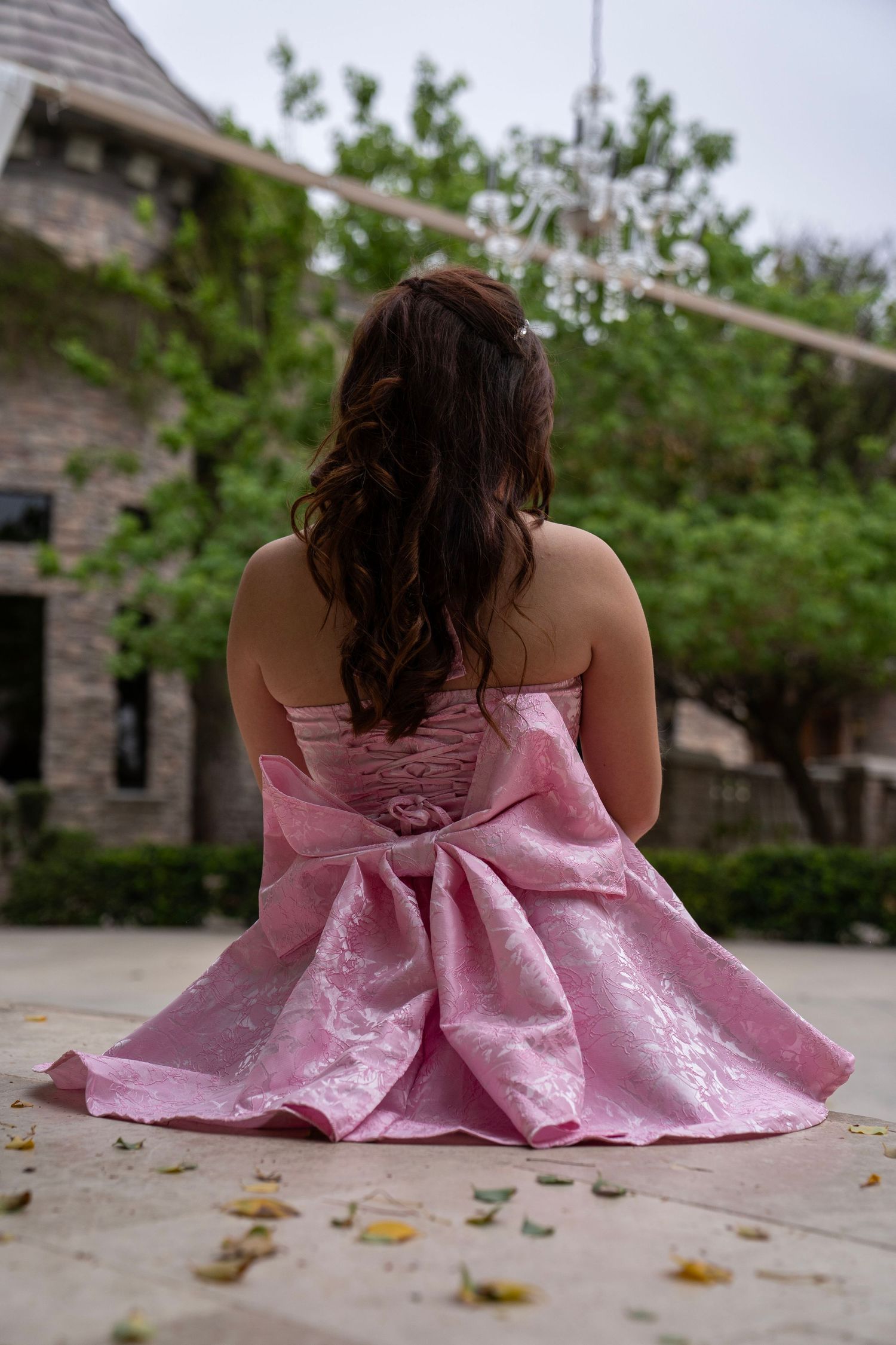 Back view of woman in strapless pink bow-back dress with long curly hair sitting outdoors near a building.