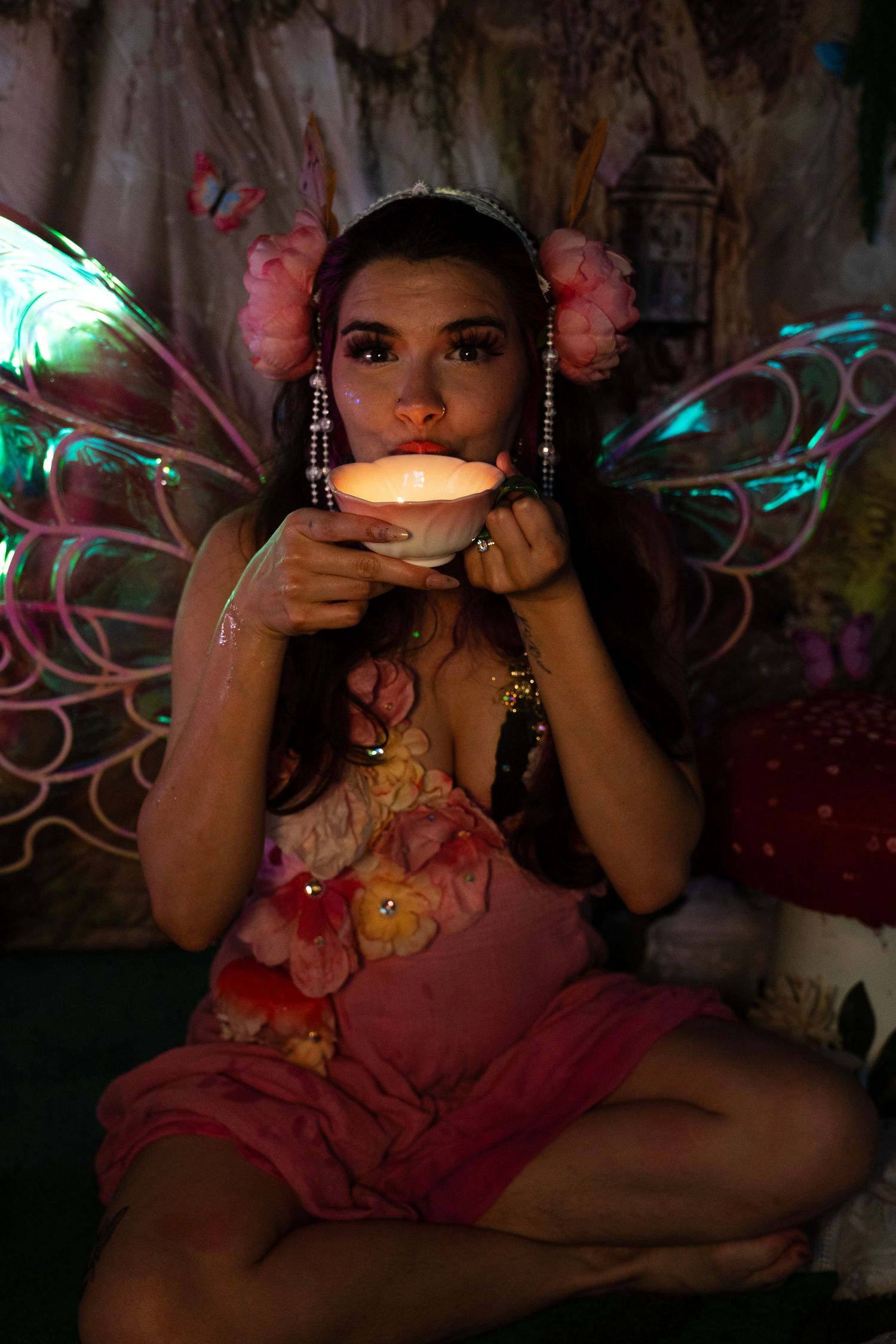 Woman in fairy costume with iridescent wings drinks from glowing lotus cup in enchanted forest scene. Fantasy Photoshoot
