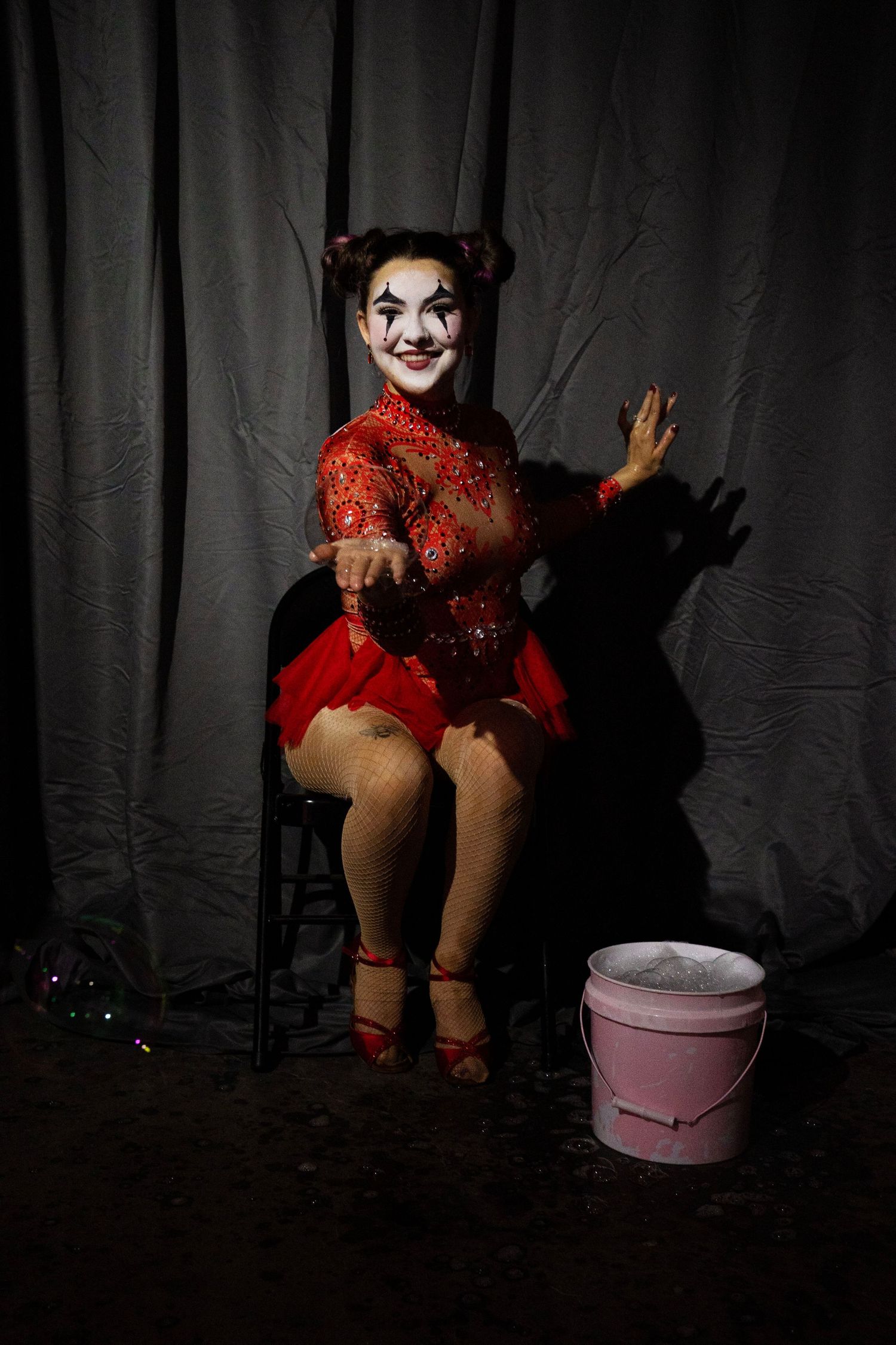 A female circus clown in red costume with white face paint sits beside a pink bucket on dark stage.