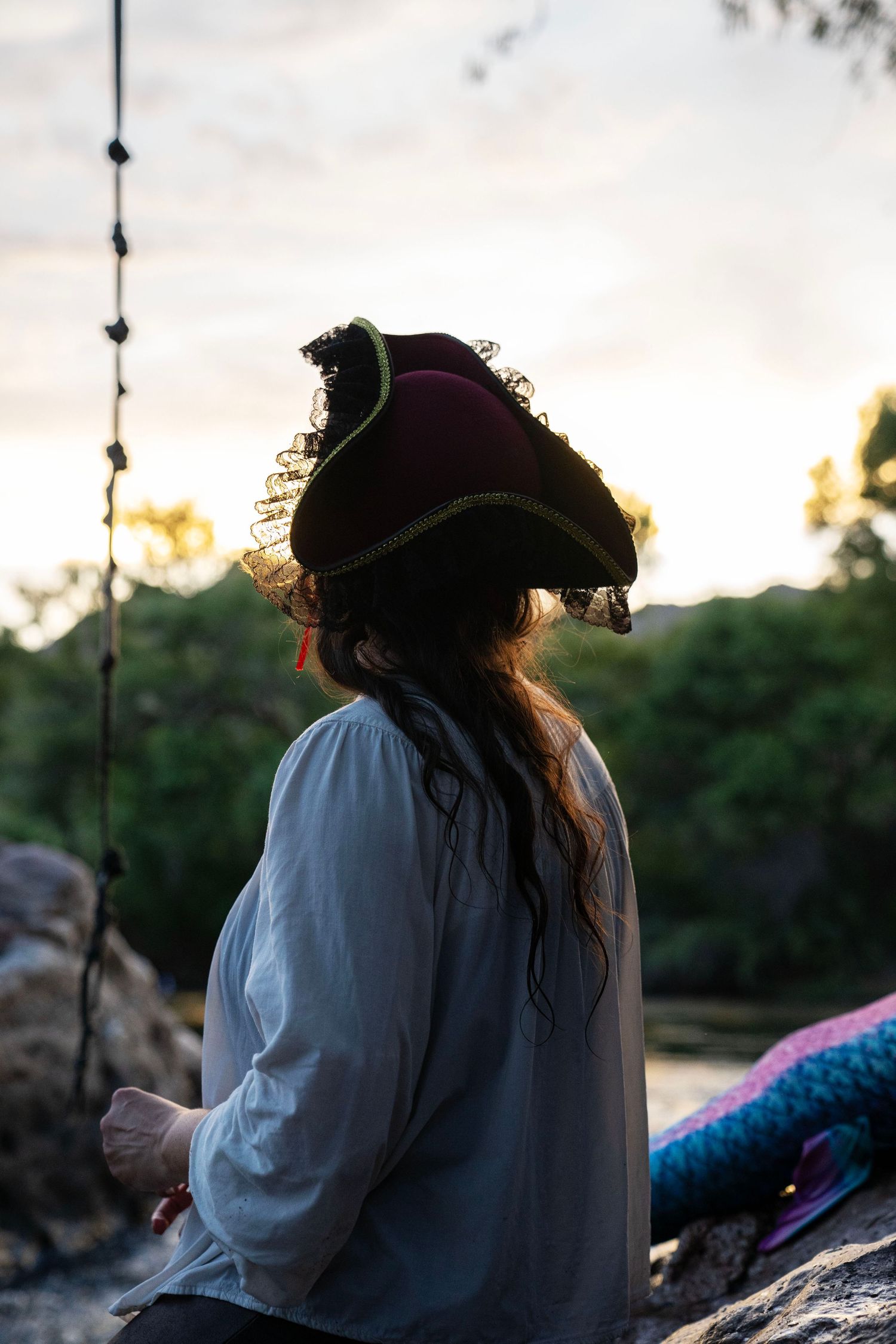 Woman in pirate hat silhouetted against golden sunset sky near colorful mermaid tail. Mermaid photoshoot, fantasy photoshoot.