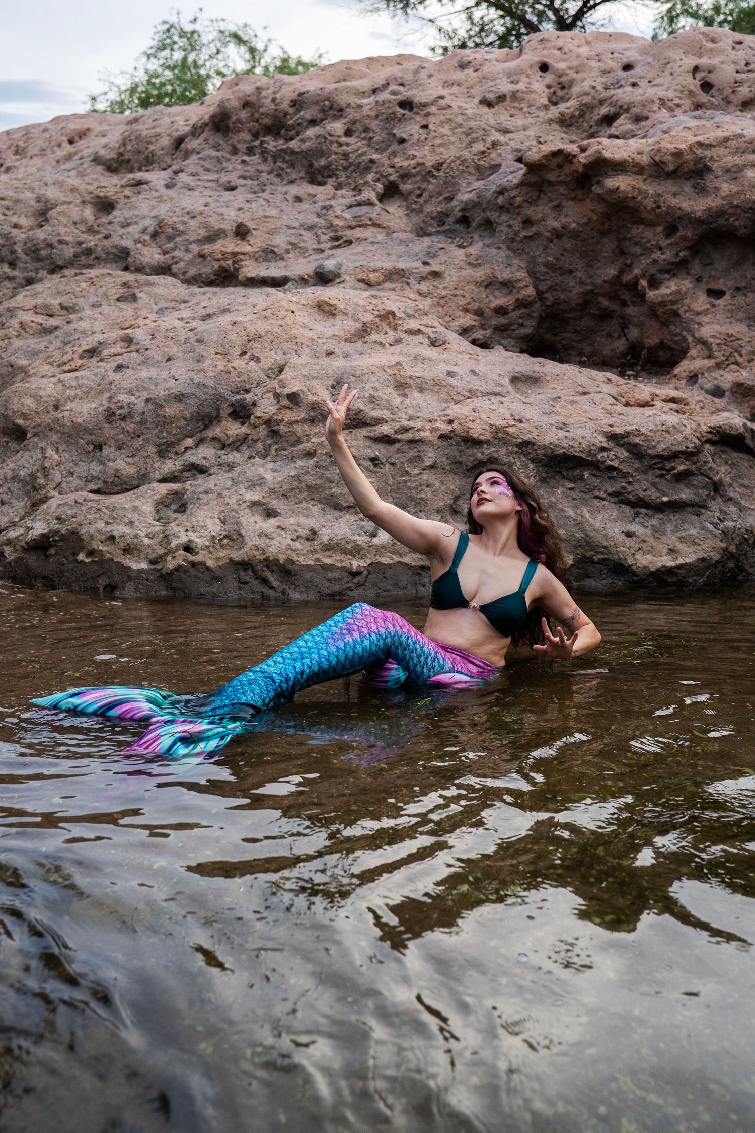 A woman in a blue and pink mermaid costume sits in water reaching upward toward rocks. Fantasy Photoshoot