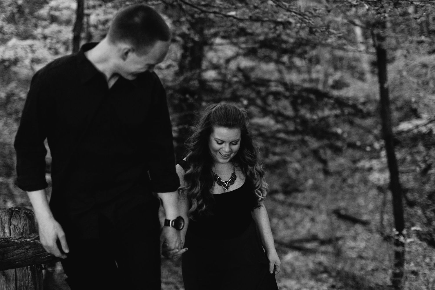 Black and white photo of couple holding hands walking through a wooded outdoor setting.