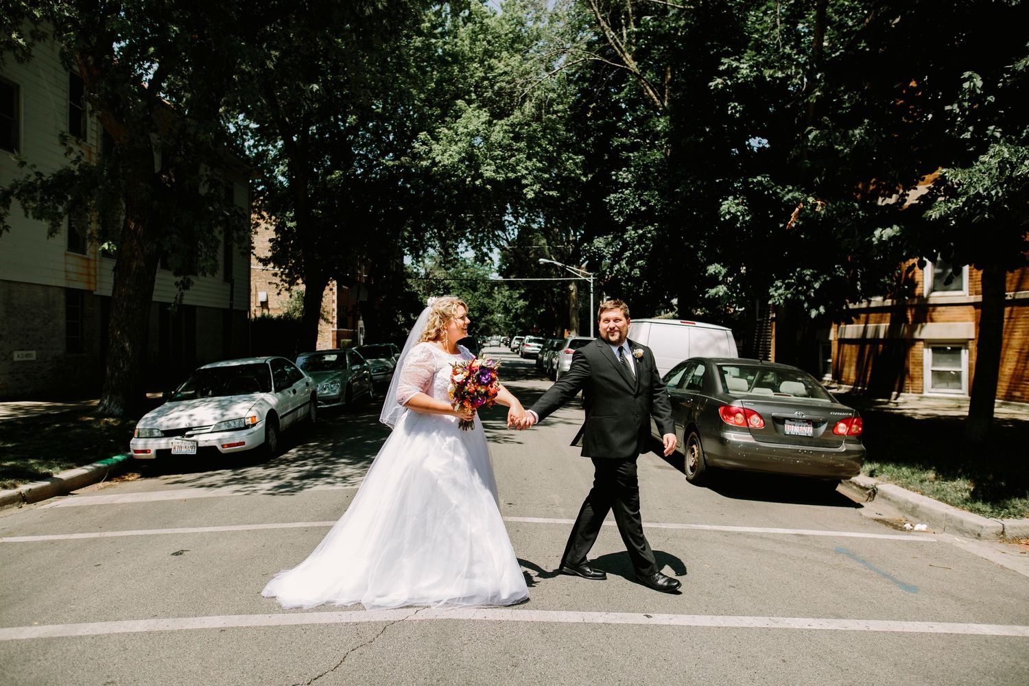 Bride and groom cross a sunny tree-lined street together on their wedding day.