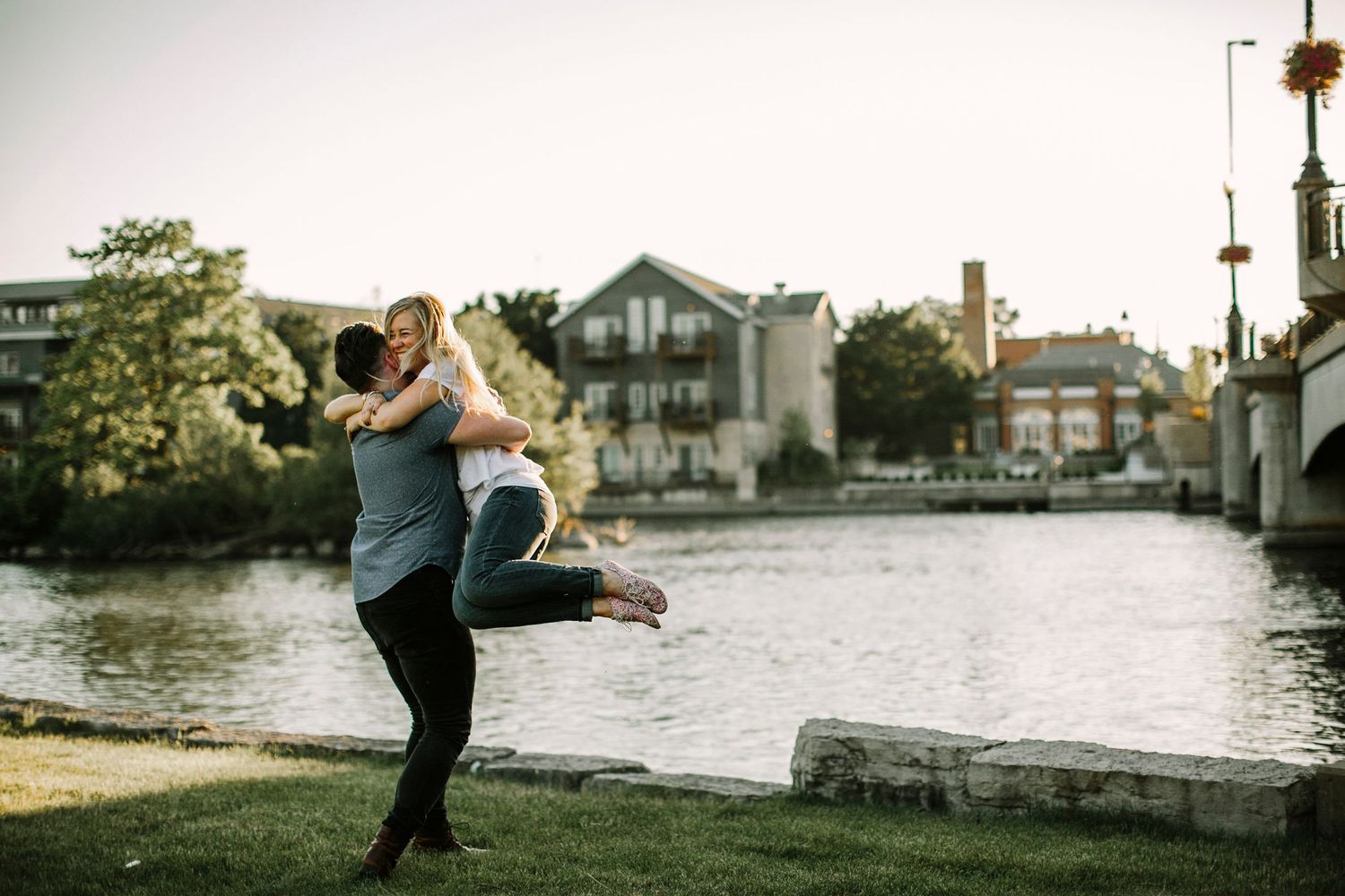 Man lifts his smiling partner joyfully by a riverside in a sunny urban park setting.