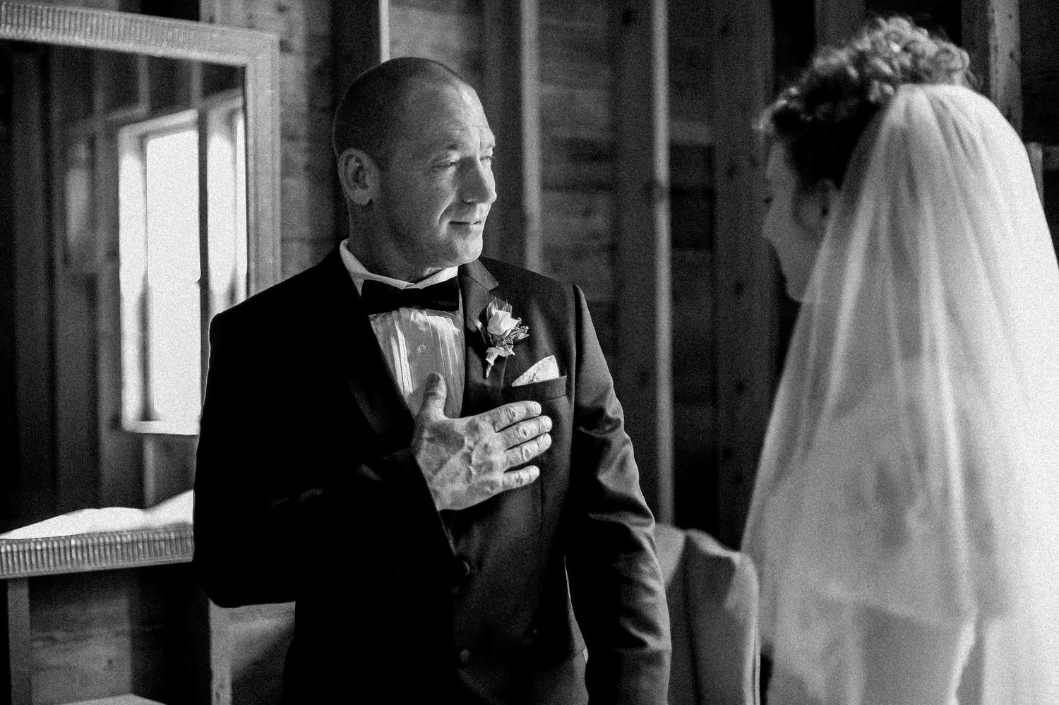 Emotional father placing hand on chest during first look with veiled bride in barn.