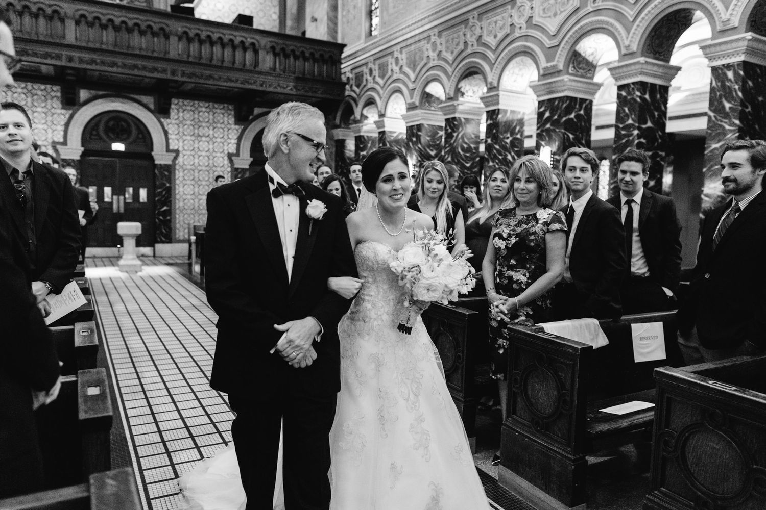 Black and white photo of bride walking down church aisle with her proud father.