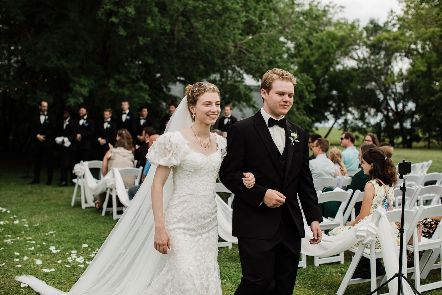 Newlyweds joyfully walking back down the aisle after outdoor wedding ceremony on green lawn.