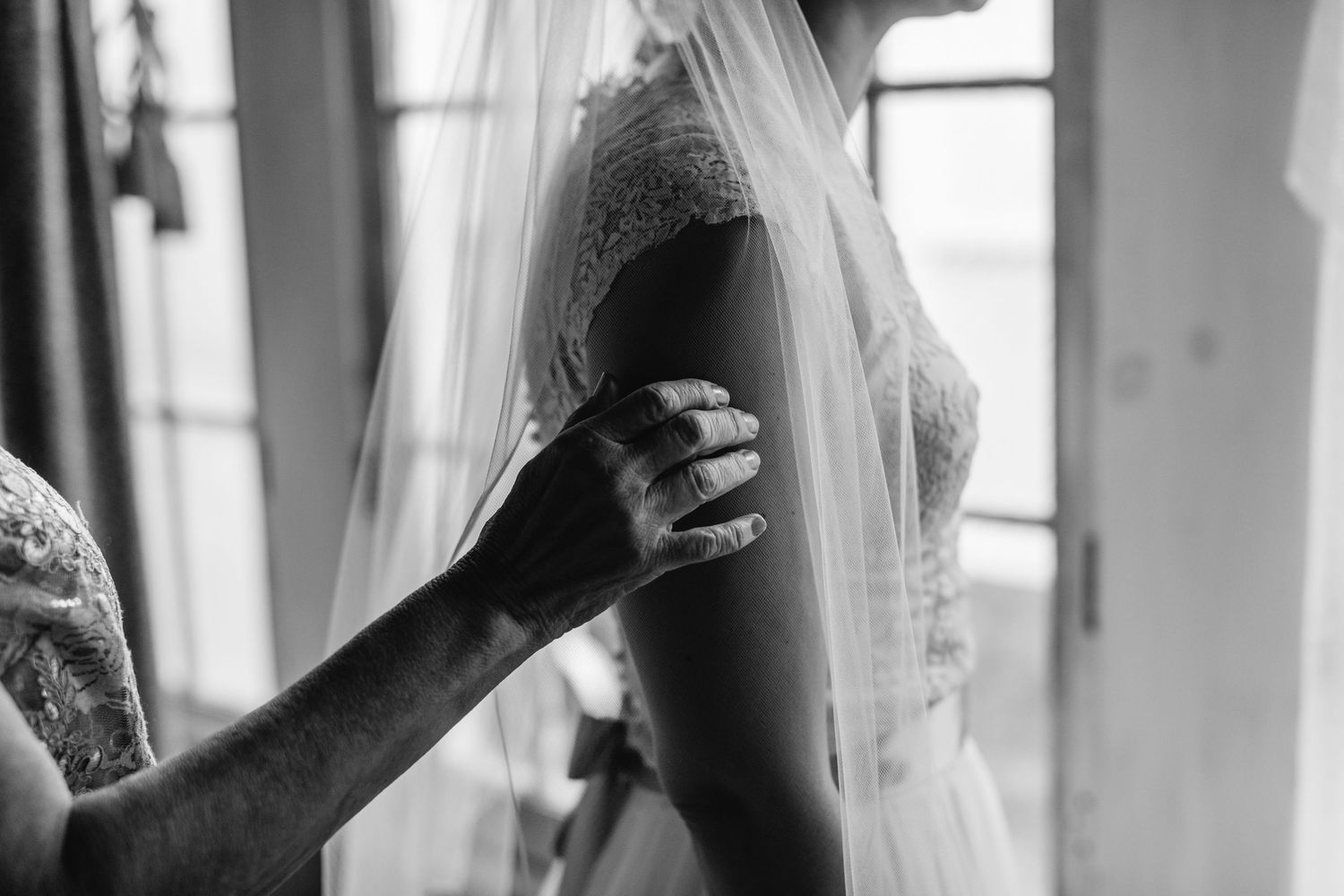 A bride in a lace wedding dress and veil being helped by a hand adjusting her gown in black and white.