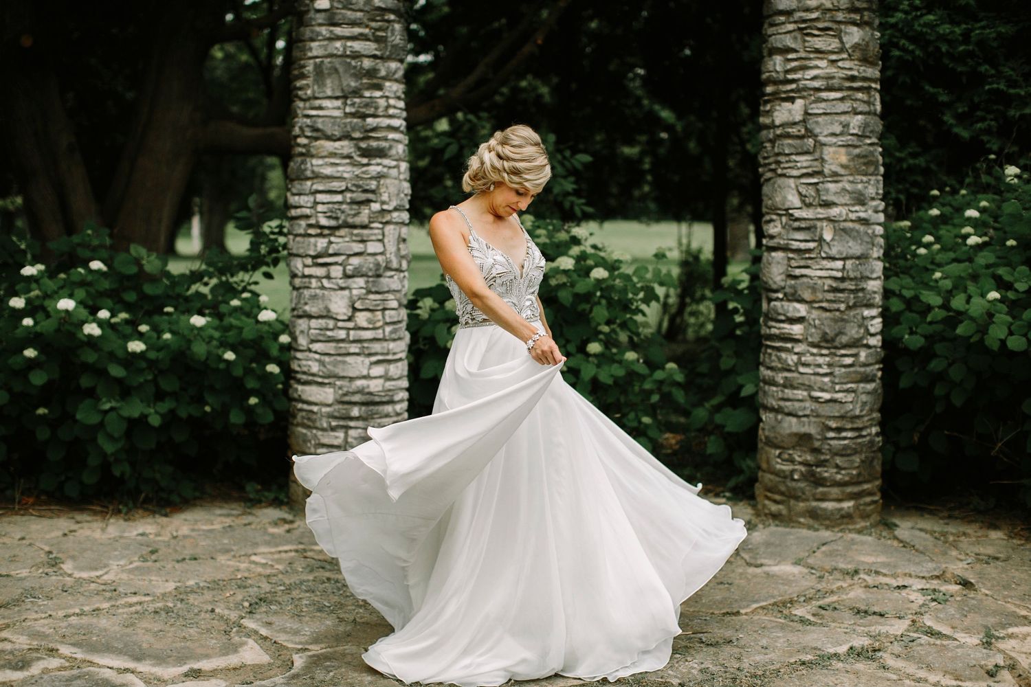 Bride in flowing white gown twirling outdoors near stone columns and lush greenery.
