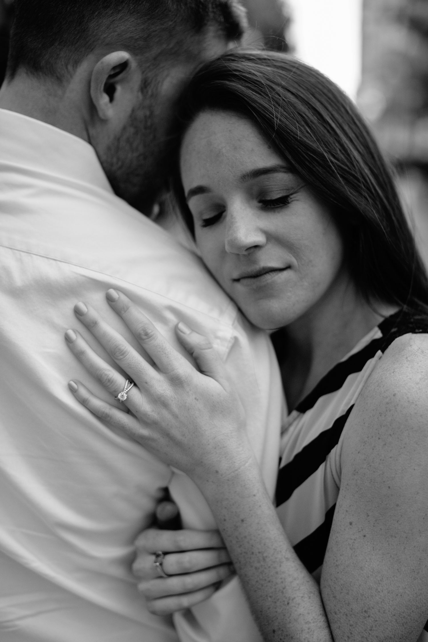 A woman with an engagement ring embraces a man, resting her head on his shoulder in a black and white photo.