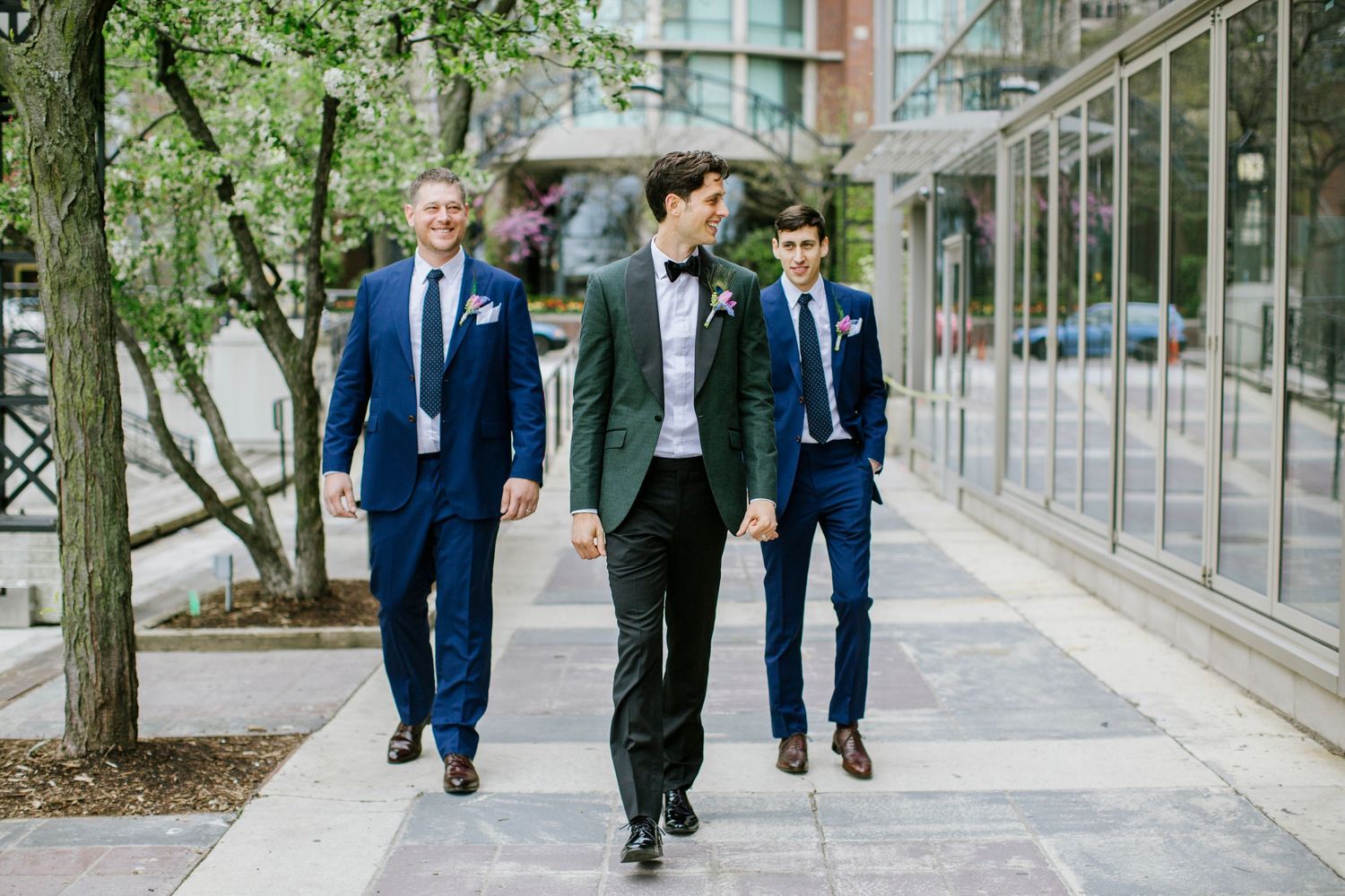 Groom in green jacket walks with two groomsmen in navy suits along urban sidewalk.