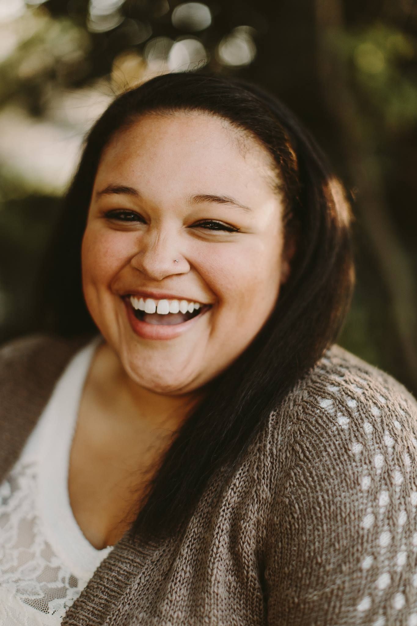 Joyful woman with dark hair laughing warmly outdoors wearing a tan knit cardigan over white top.