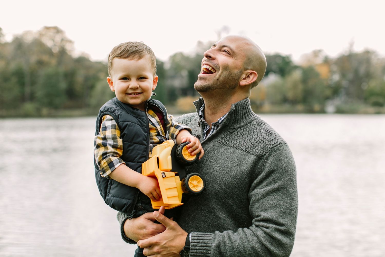 Happy father holding smiling toddler with yellow toy truck near a scenic lakeside.