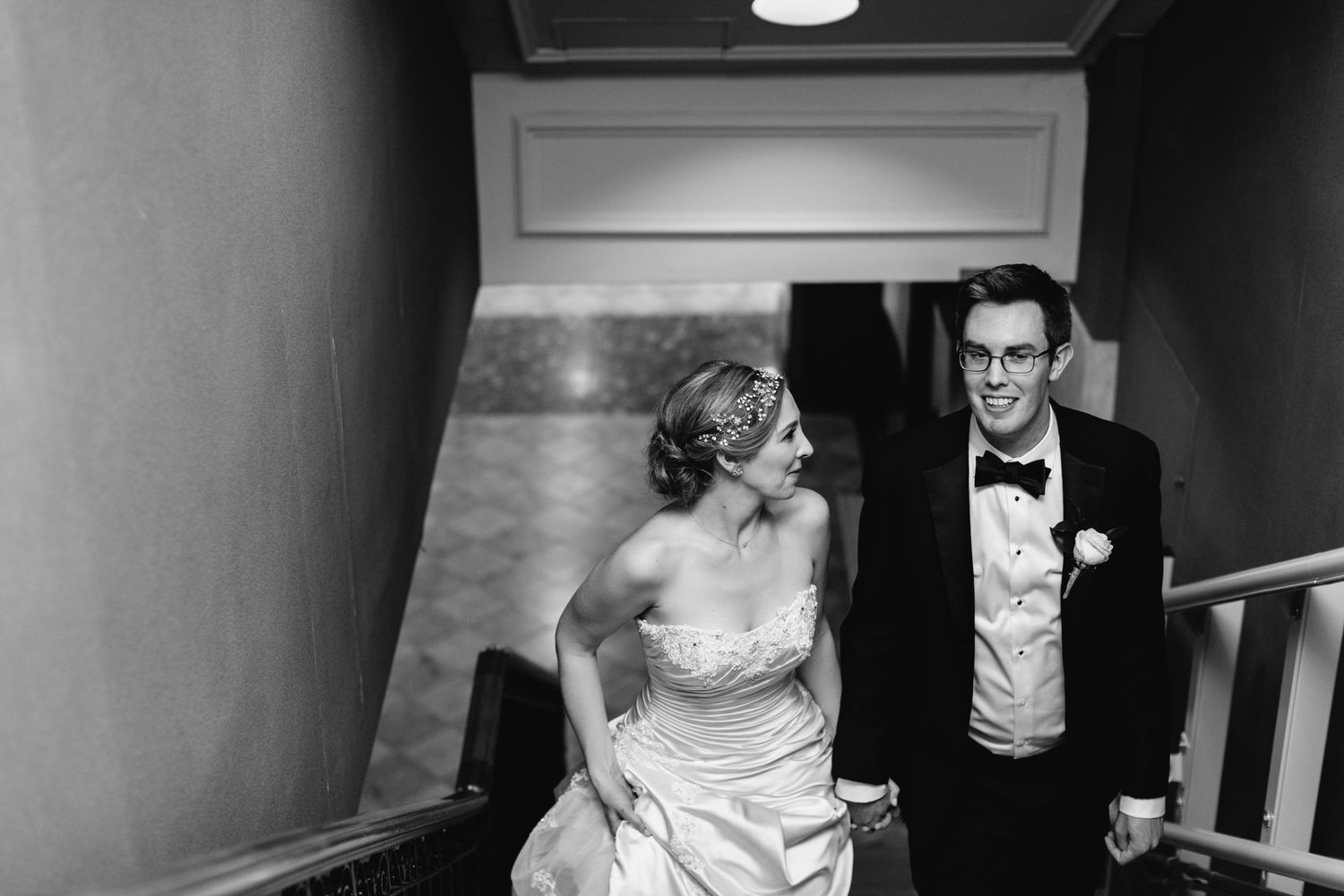 Black and white photo of smiling bride and groom descending an elegant staircase together.