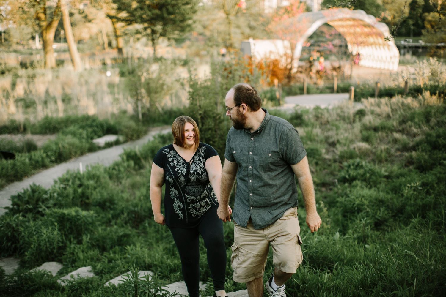 Couple holding hands and smiling while walking through lush garden path at golden hour.