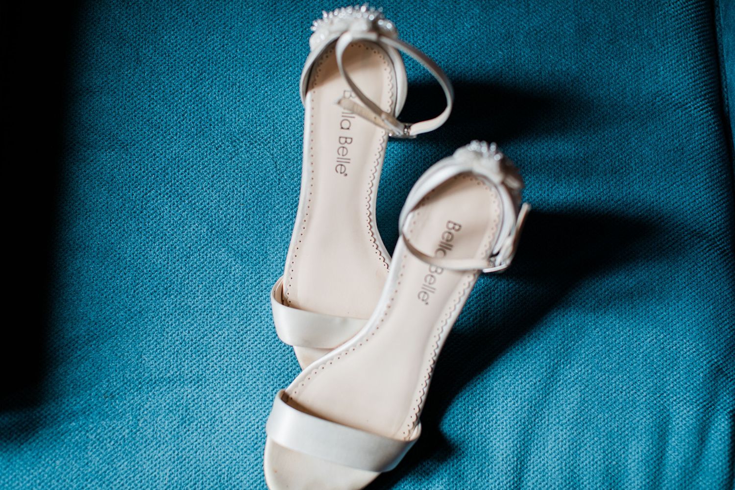 Elegant white bridal heeled sandals with ankle strap displayed on teal blue chair.
