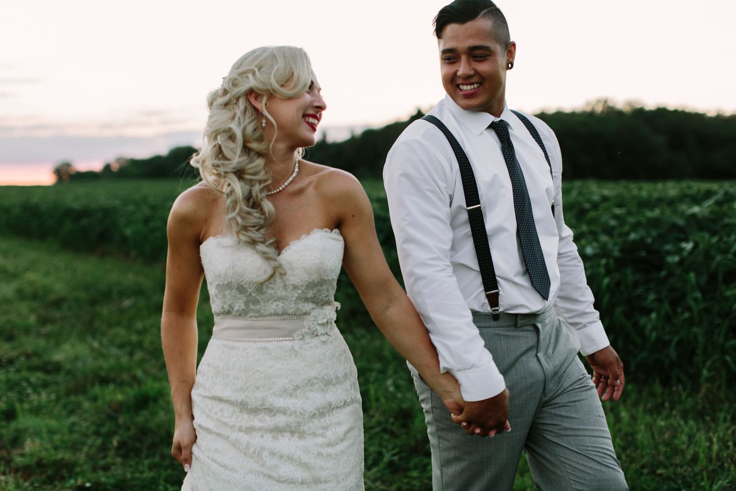 Happy bride and groom holding hands walking through green field at sunset.