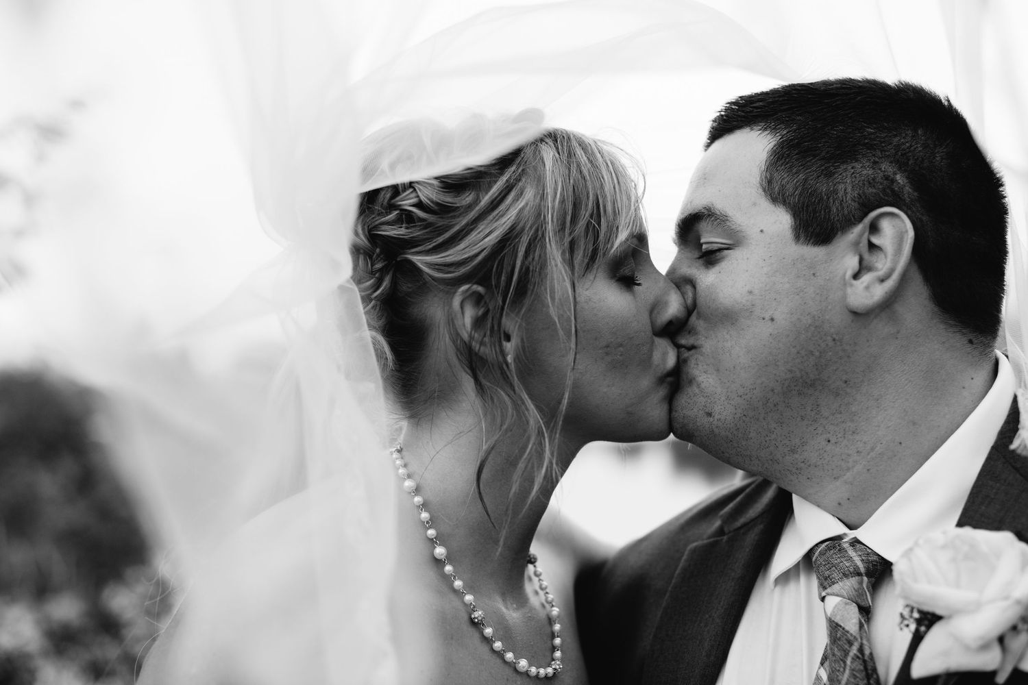 Bride and groom sharing romantic kiss under flowing veil in black and white portrait.