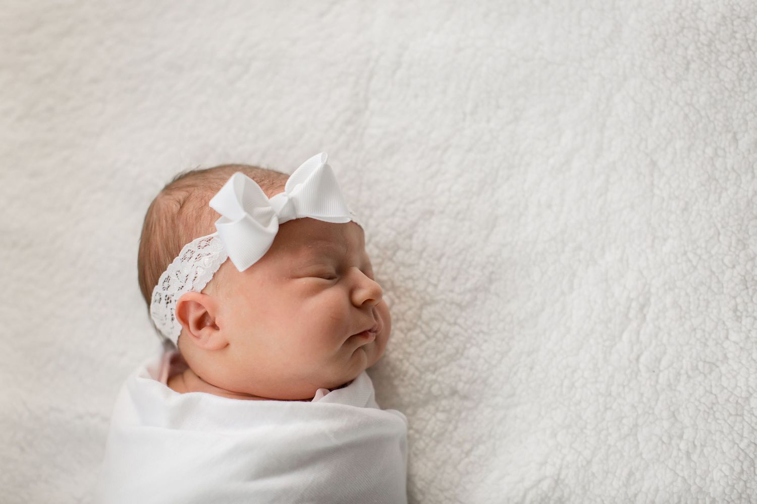 Peaceful newborn baby girl swaddled in white with a delicate bow headband sleeping.