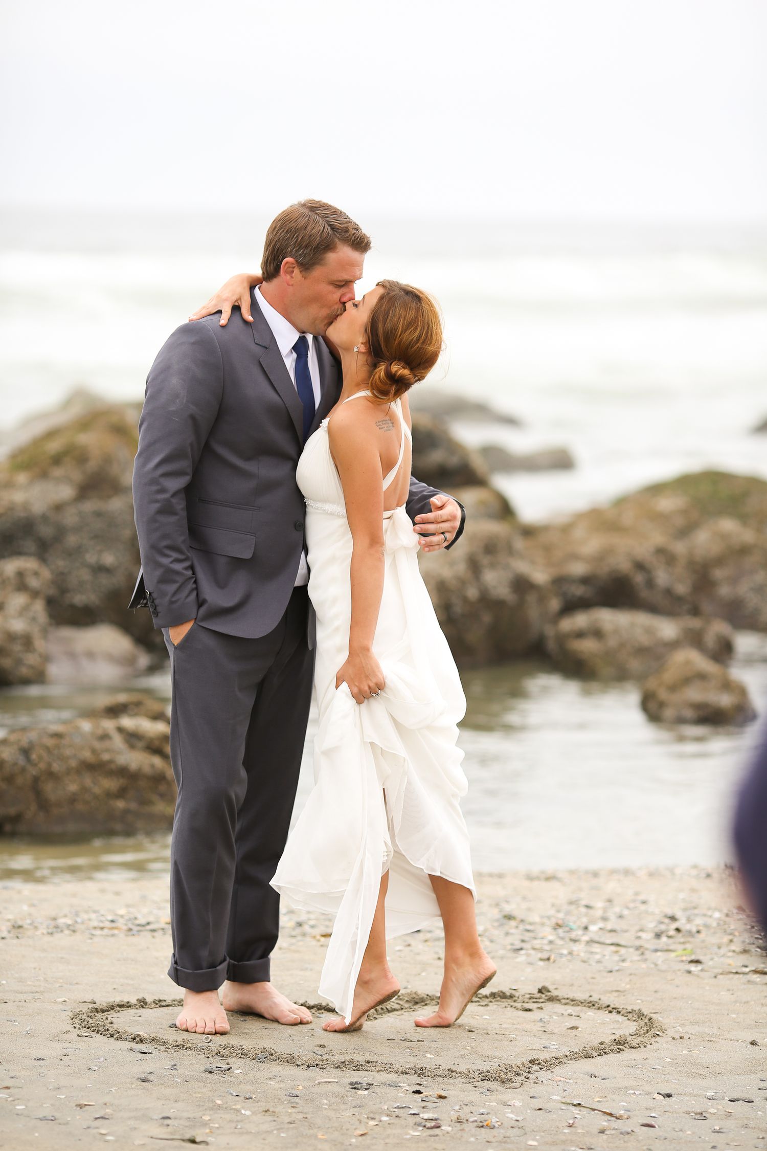 Bride and groom share a barefoot kiss inside a heart drawn in beach sand by the ocean.