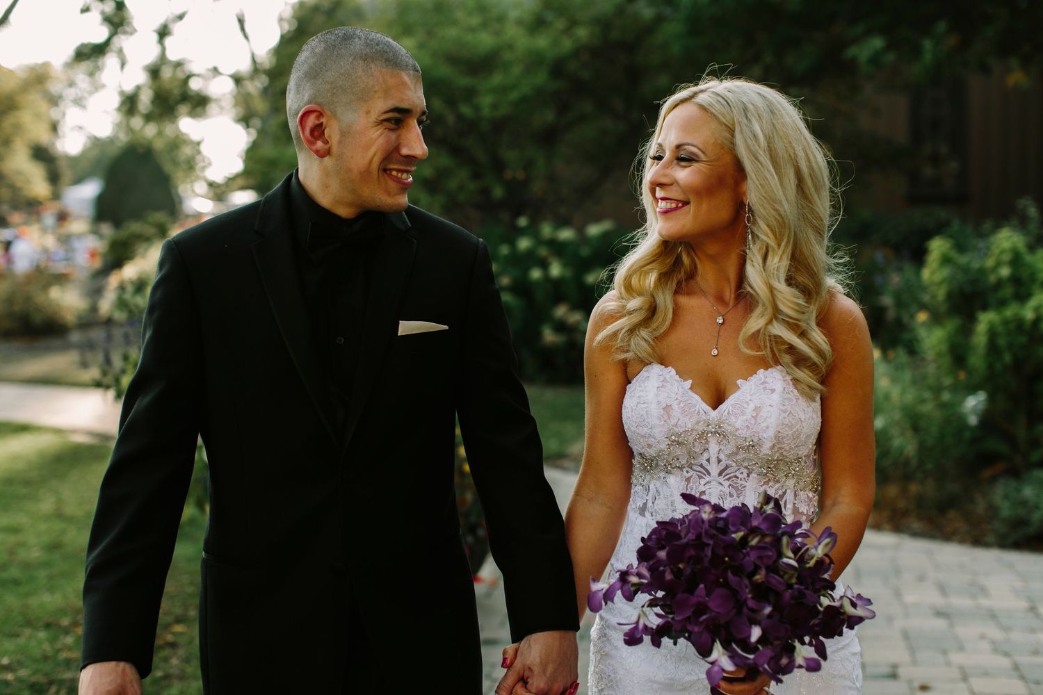Smiling bride in lace gown holding purple bouquet walks with groom in black suit.