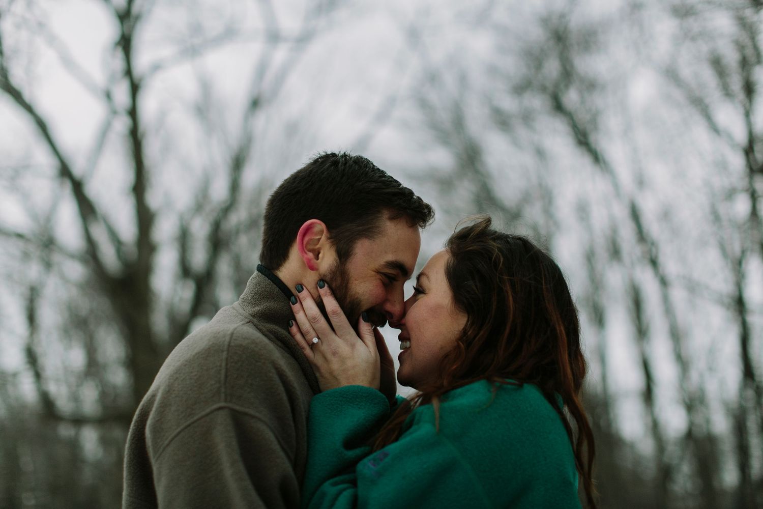 An engaged couple shares a tender nose-to-nose moment with bare winter trees in the background.