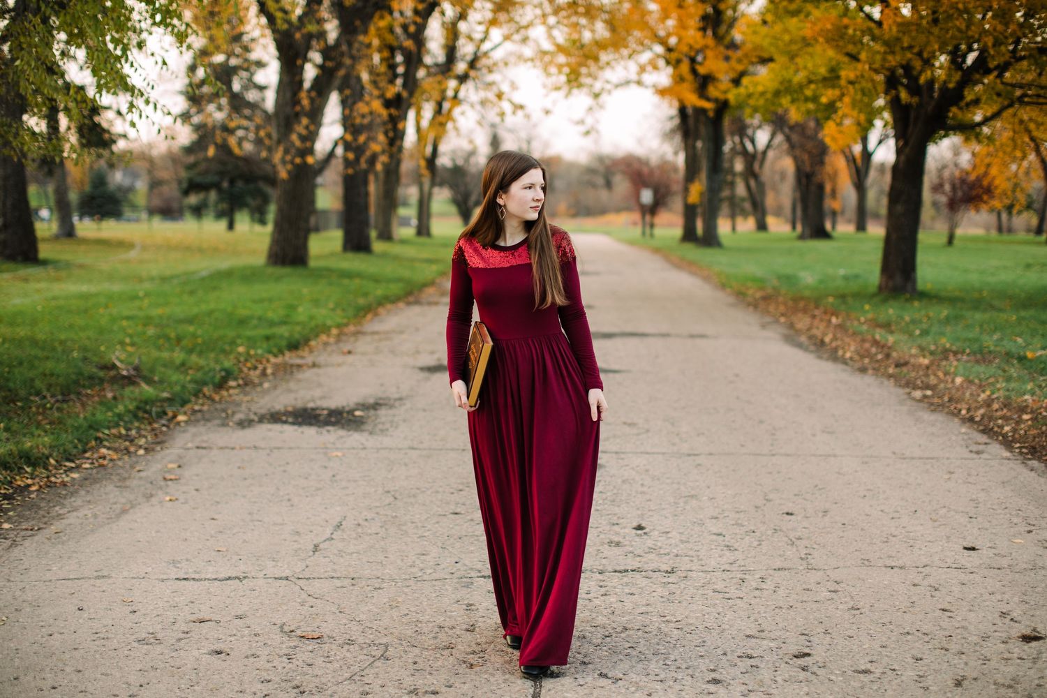 Woman in elegant burgundy maxi dress walking along tree-lined path during autumn fall season.