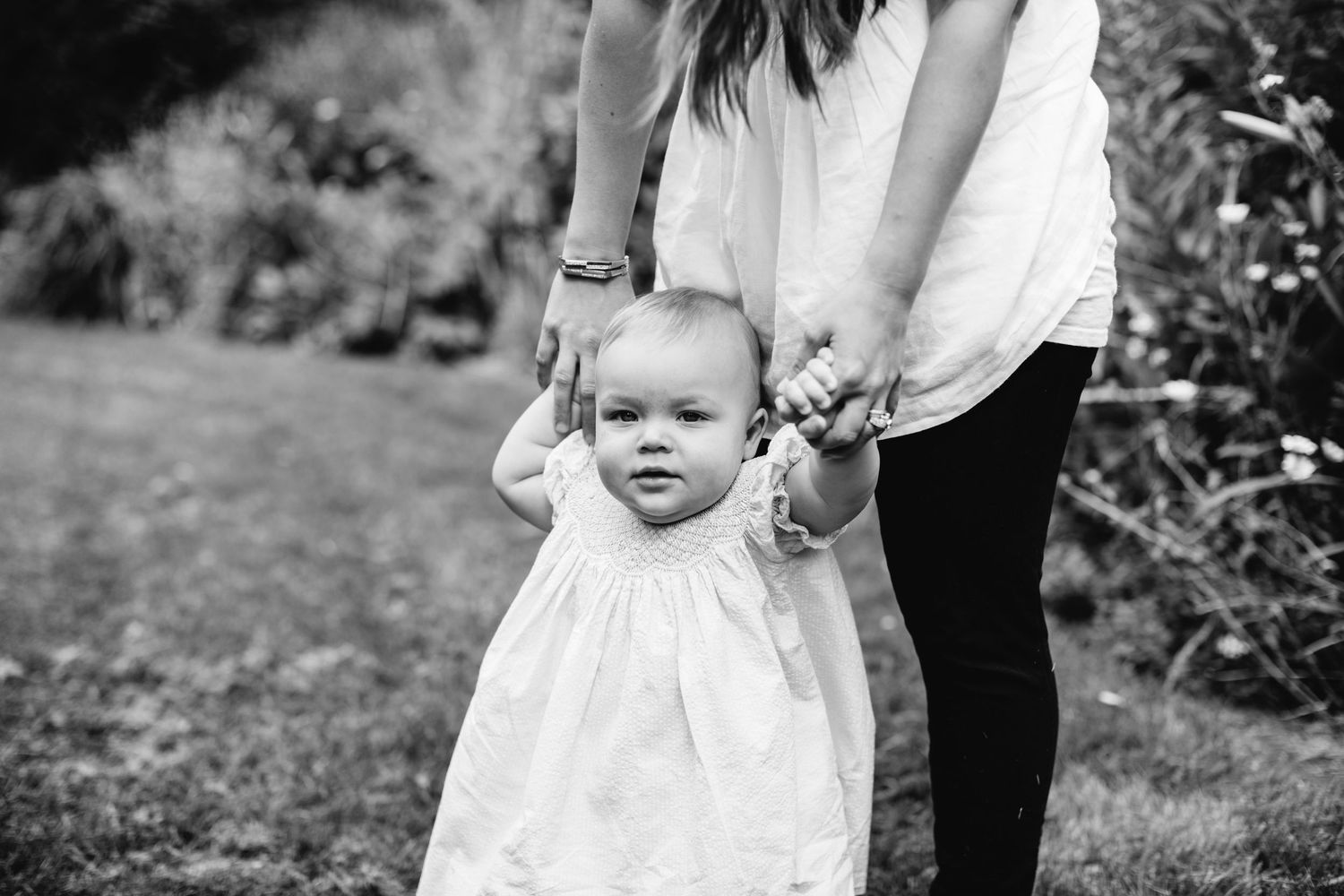 Black and white photo of baby in white dress learning to walk with mothers help outdoors.