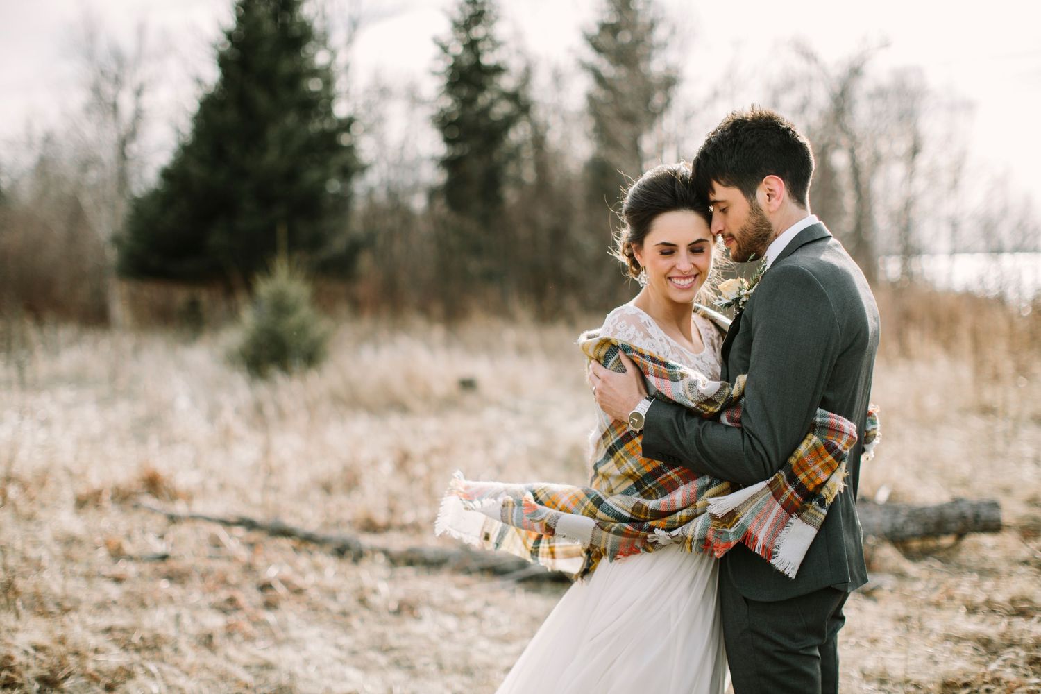 Groom wraps bride in a plaid blanket as they embrace in a wintry outdoor field.