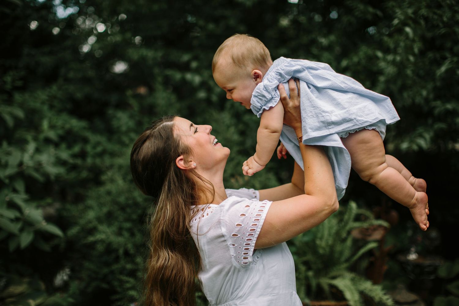 Joyful mother lifting smiling baby in blue dress outdoors surrounded by lush greenery.
