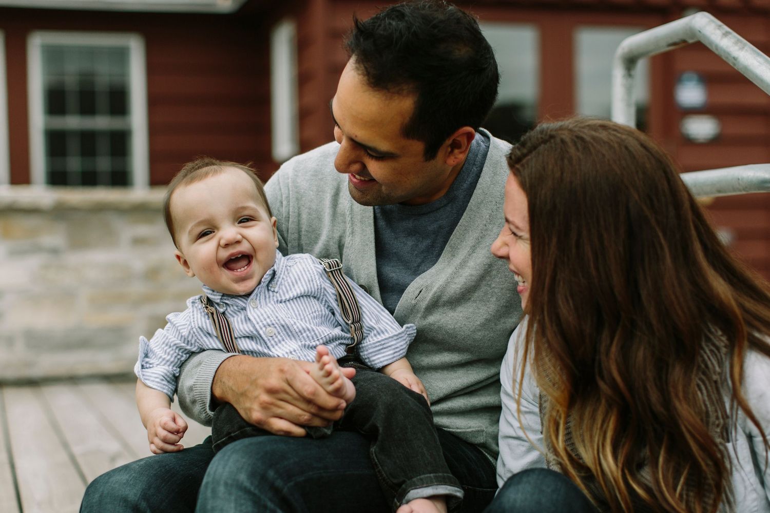 Joyful family with laughing baby boy sitting together on wooden steps near red barn.