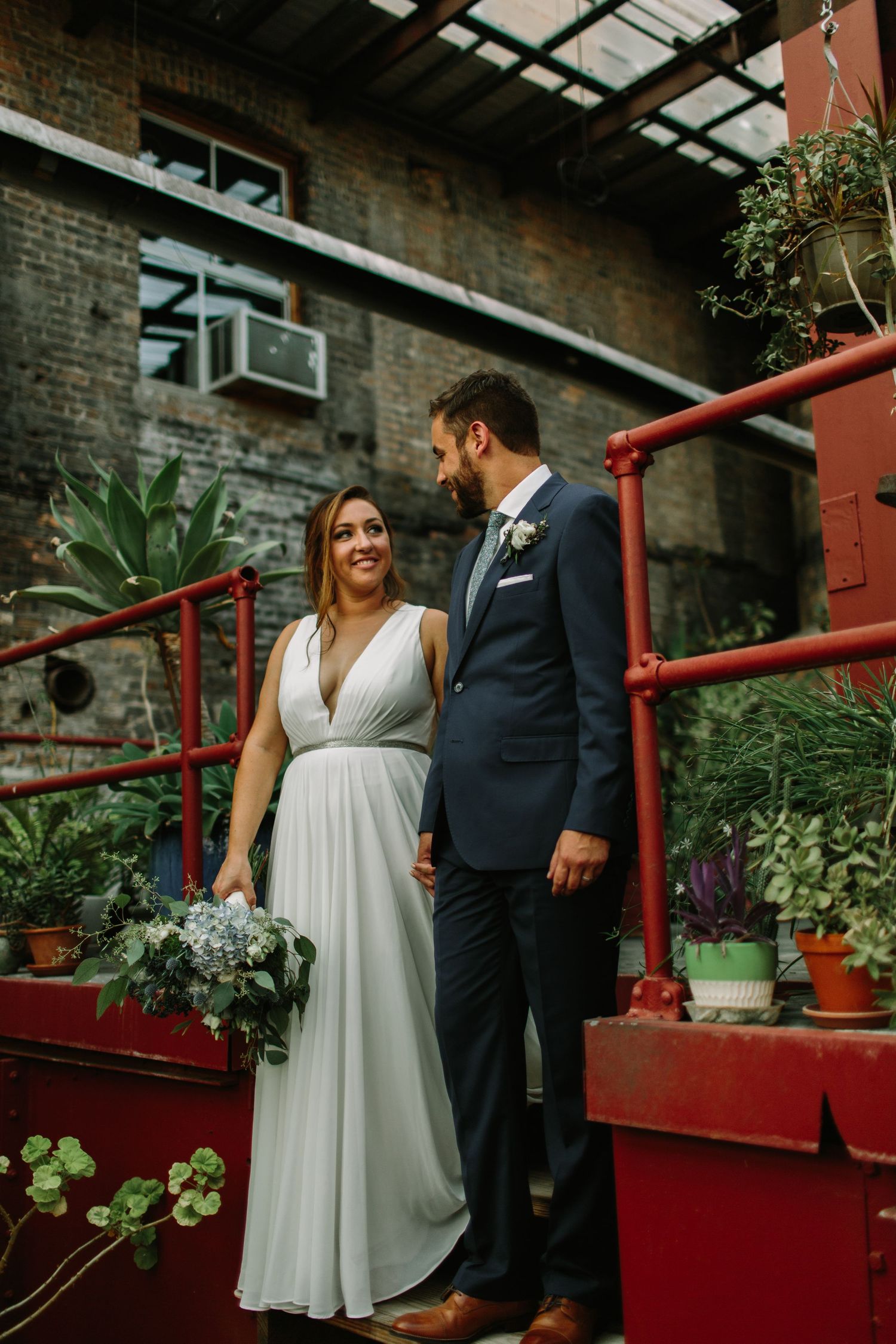 Couple in wedding attire holding hands on red metal stairs surrounded by potted plants.