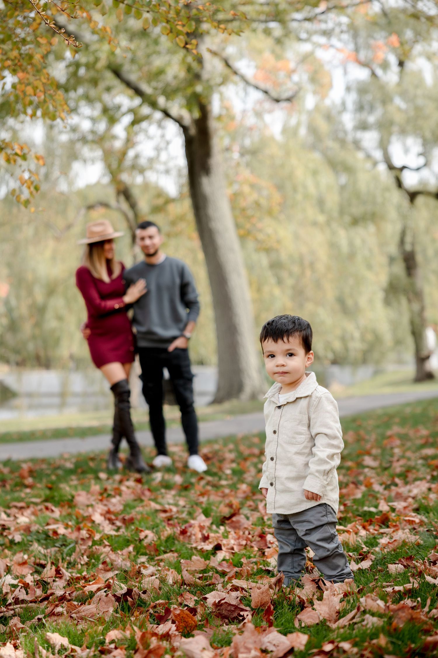 Toddler standing in autumn leaves with parents in background.