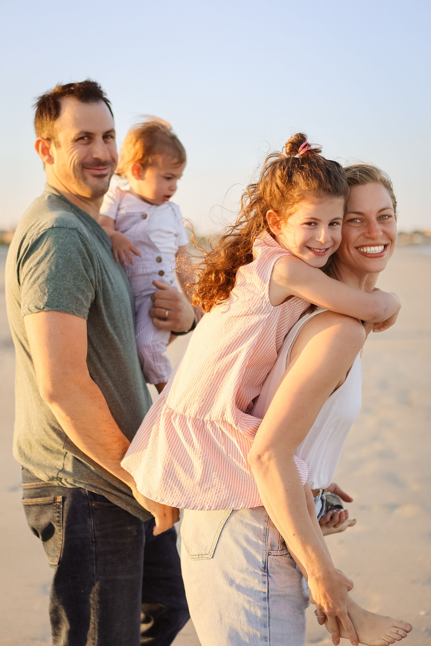 Happy white family of four enjoying sunset beach walk, mother giving daughter piggyback ride.