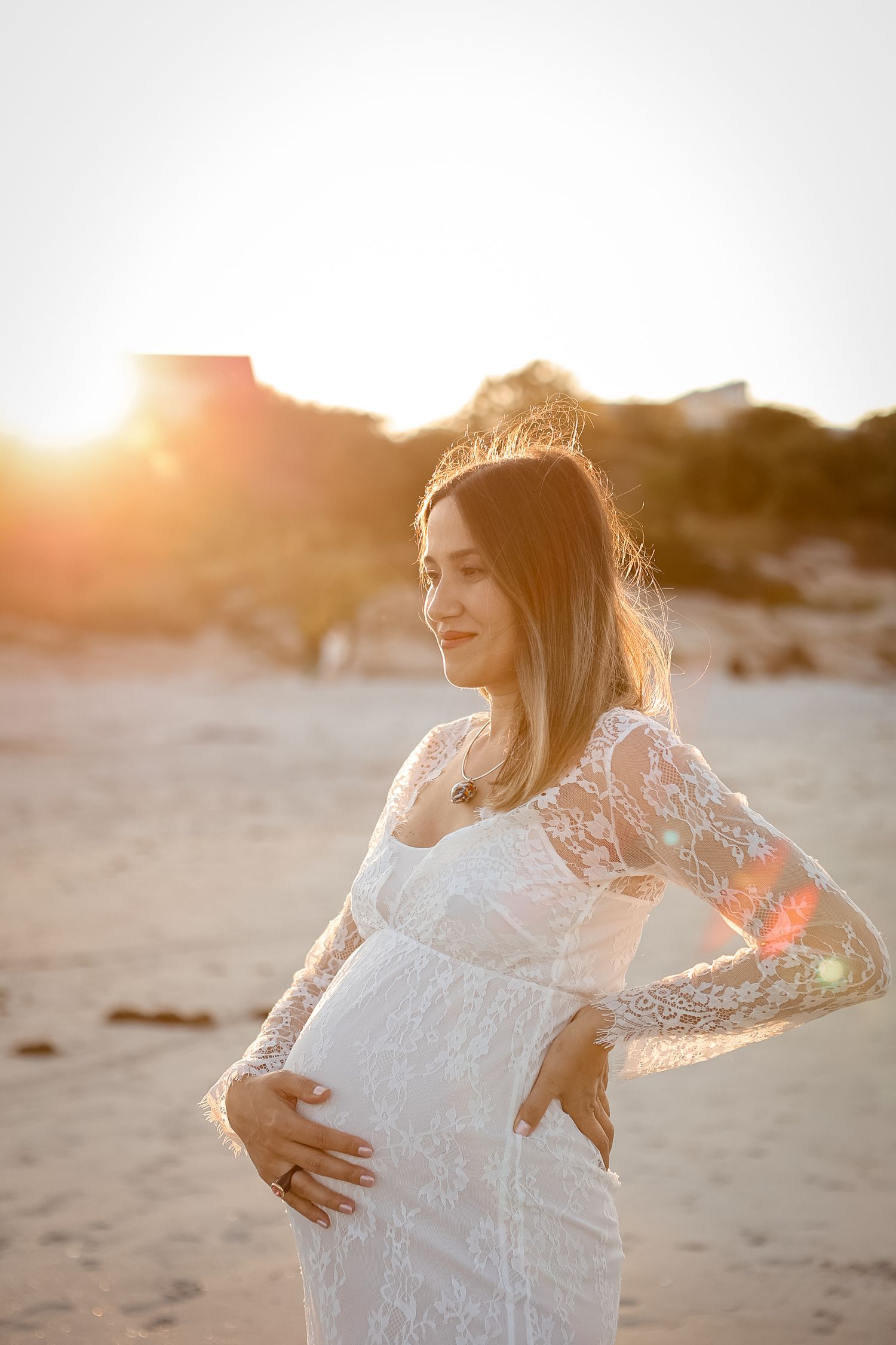 Pregnant woman in white lace dress holding baby bump on beach during golden sunset.
