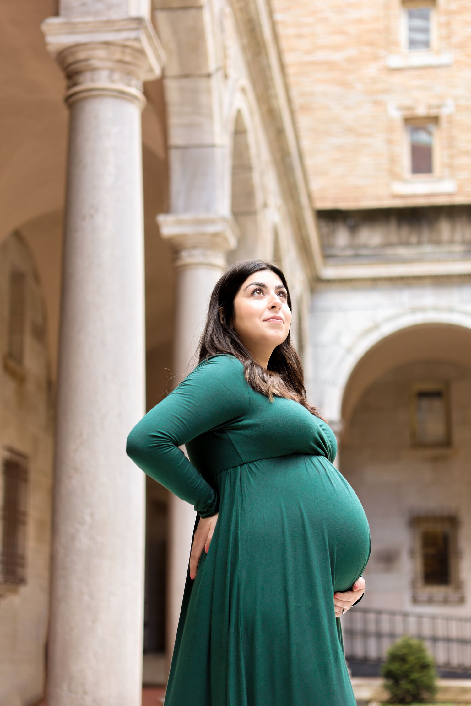 Pregnant woman in green maxi dress looking up gracefully while holding her baby bump near stone columns.