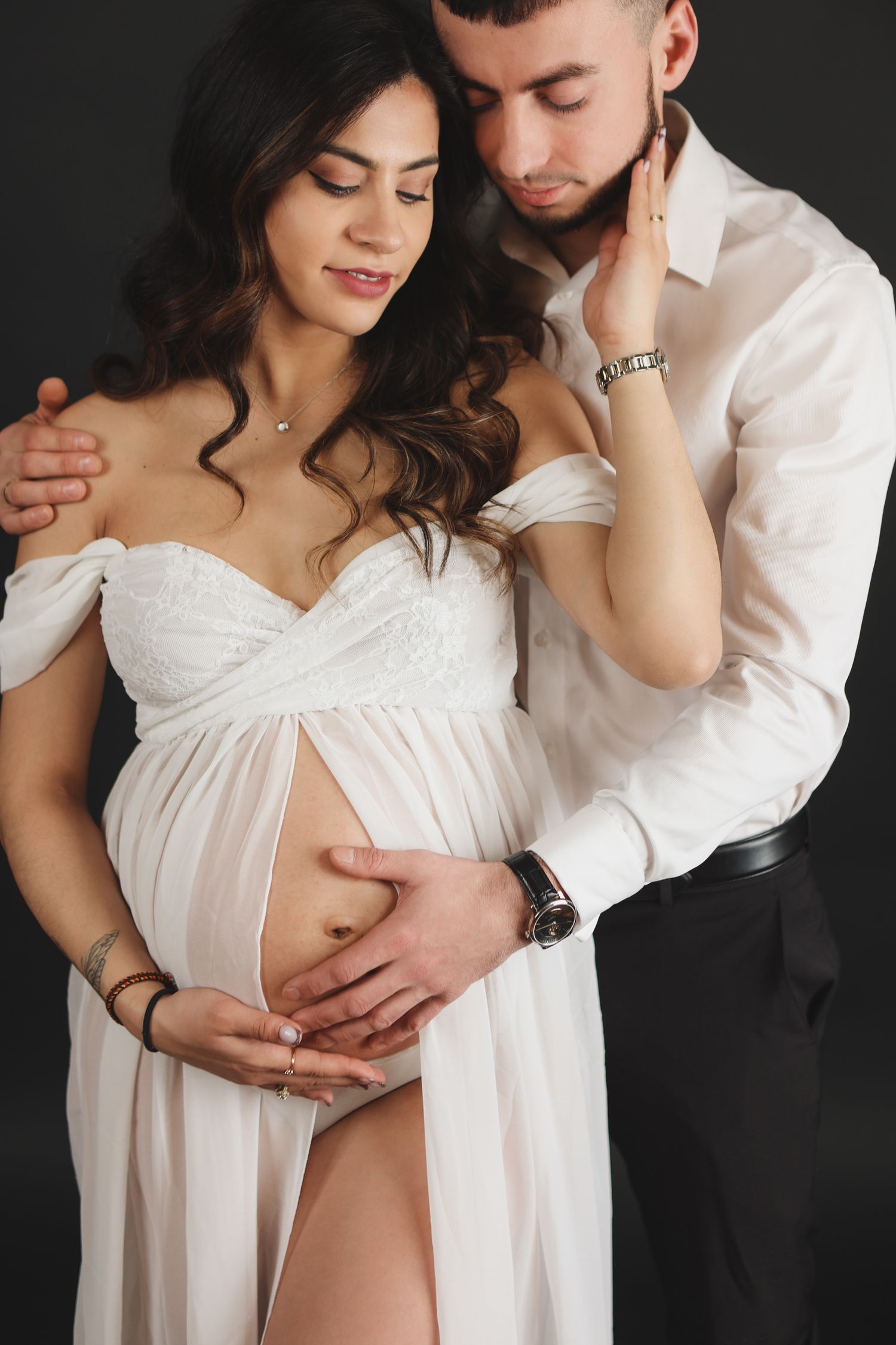 Pregnant woman in white dress with partner tenderly holding her baby bump in dark studio.