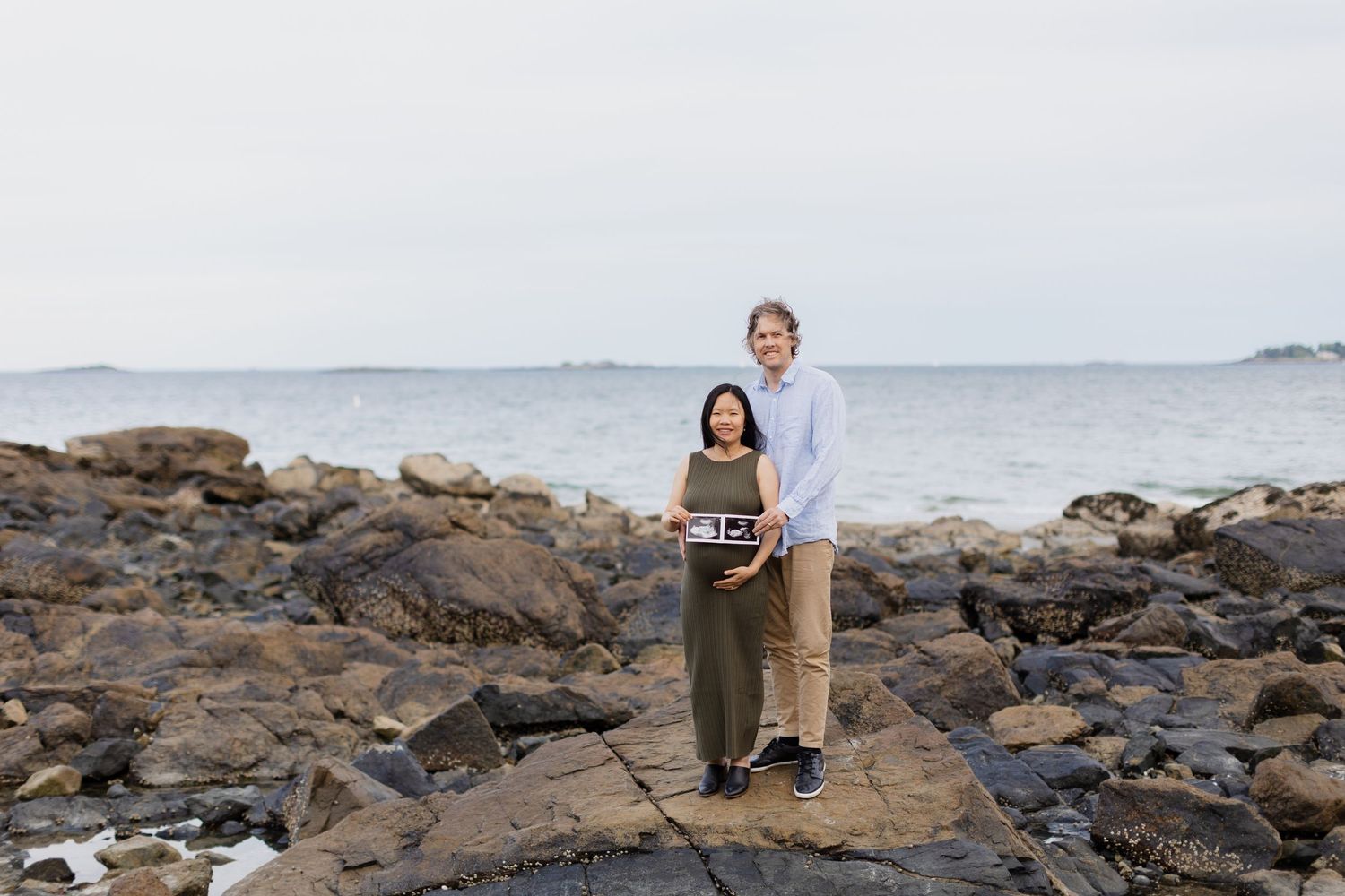 Couple standing on rocky shoreline holding ultrasound photos during an outdoor coastal maternity photoshoot.
