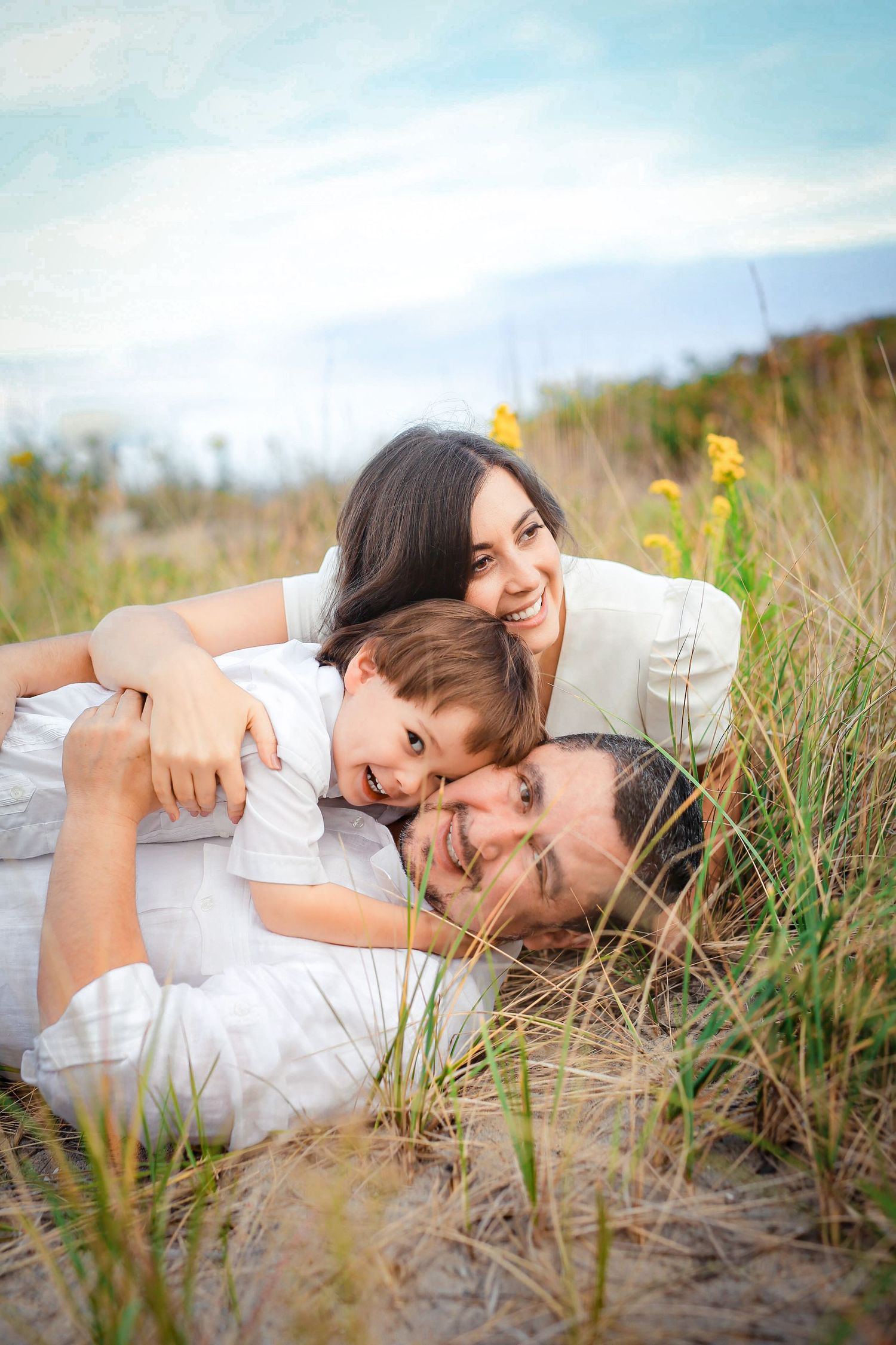 Joyful family of three lying in tall grass on a sandy beach, smiling and embracing.