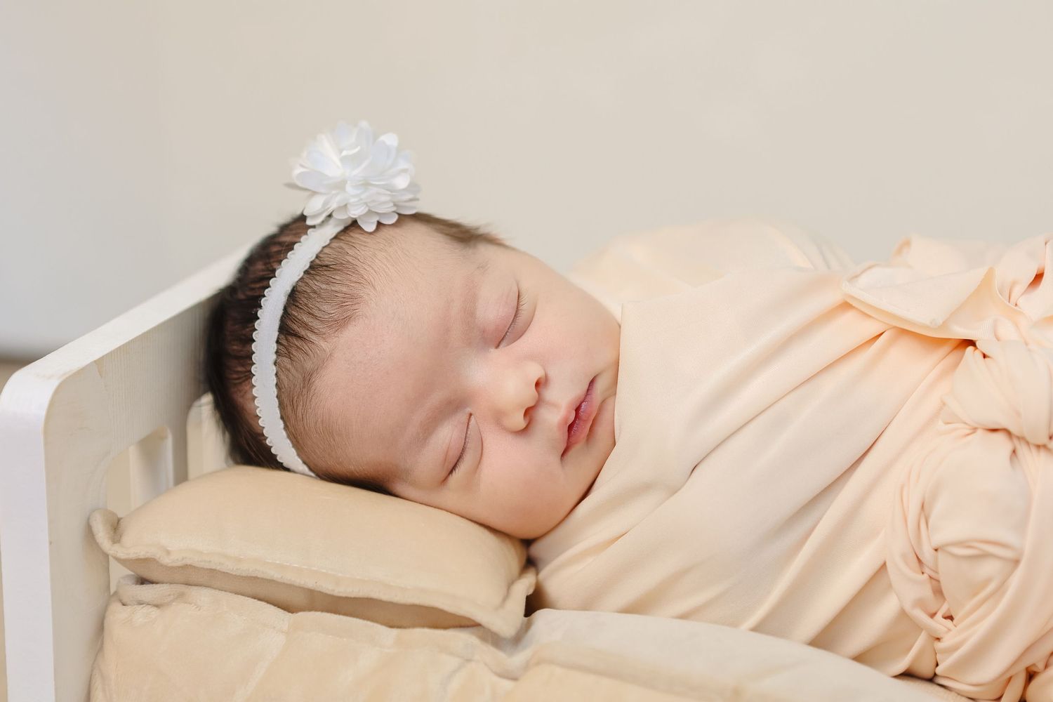 Sleeping newborn baby in peach swaddle with white flower headband in crib.