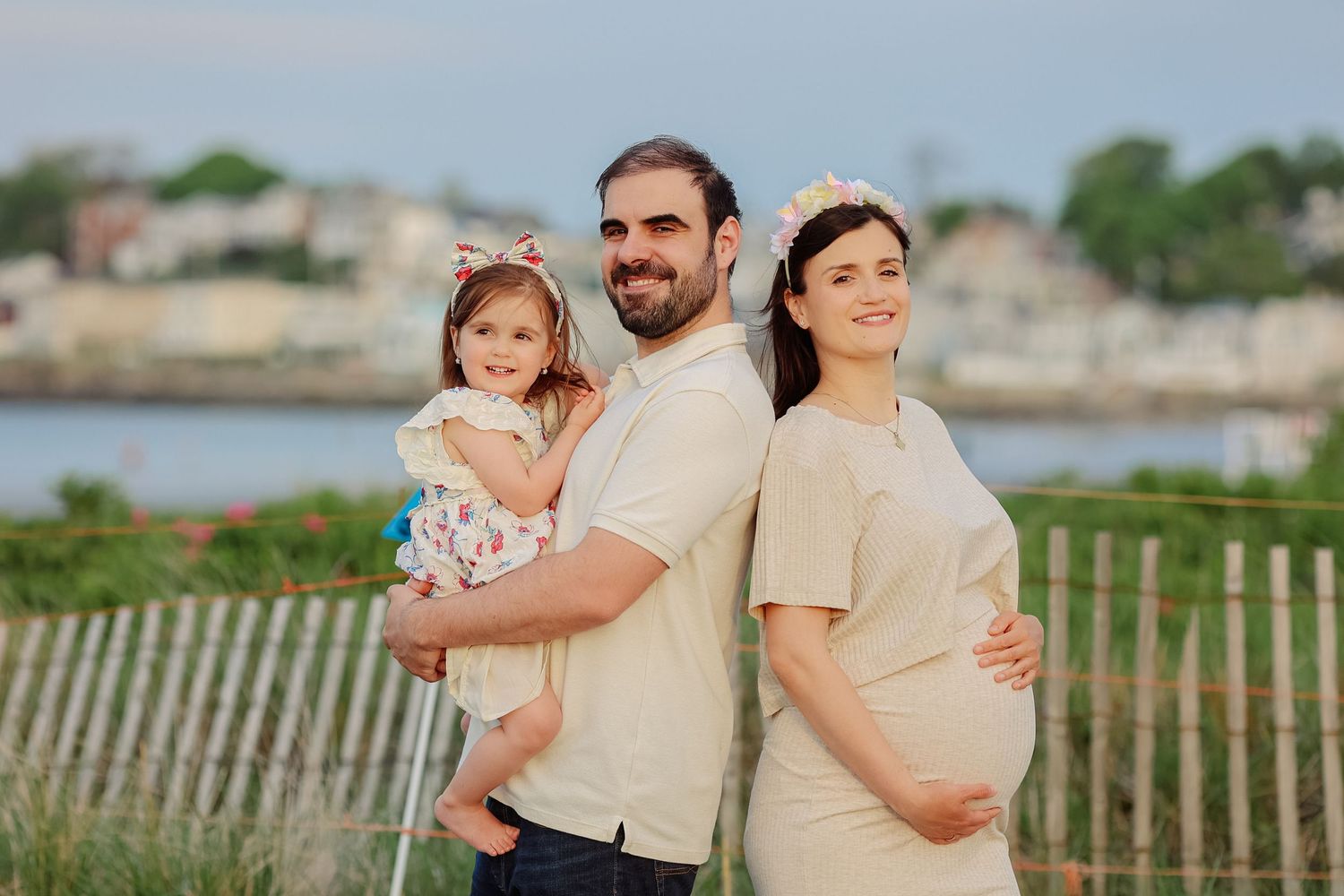 Happy family with pregnant mother, bearded father, and toddler daughter pose together near a waterfront.
