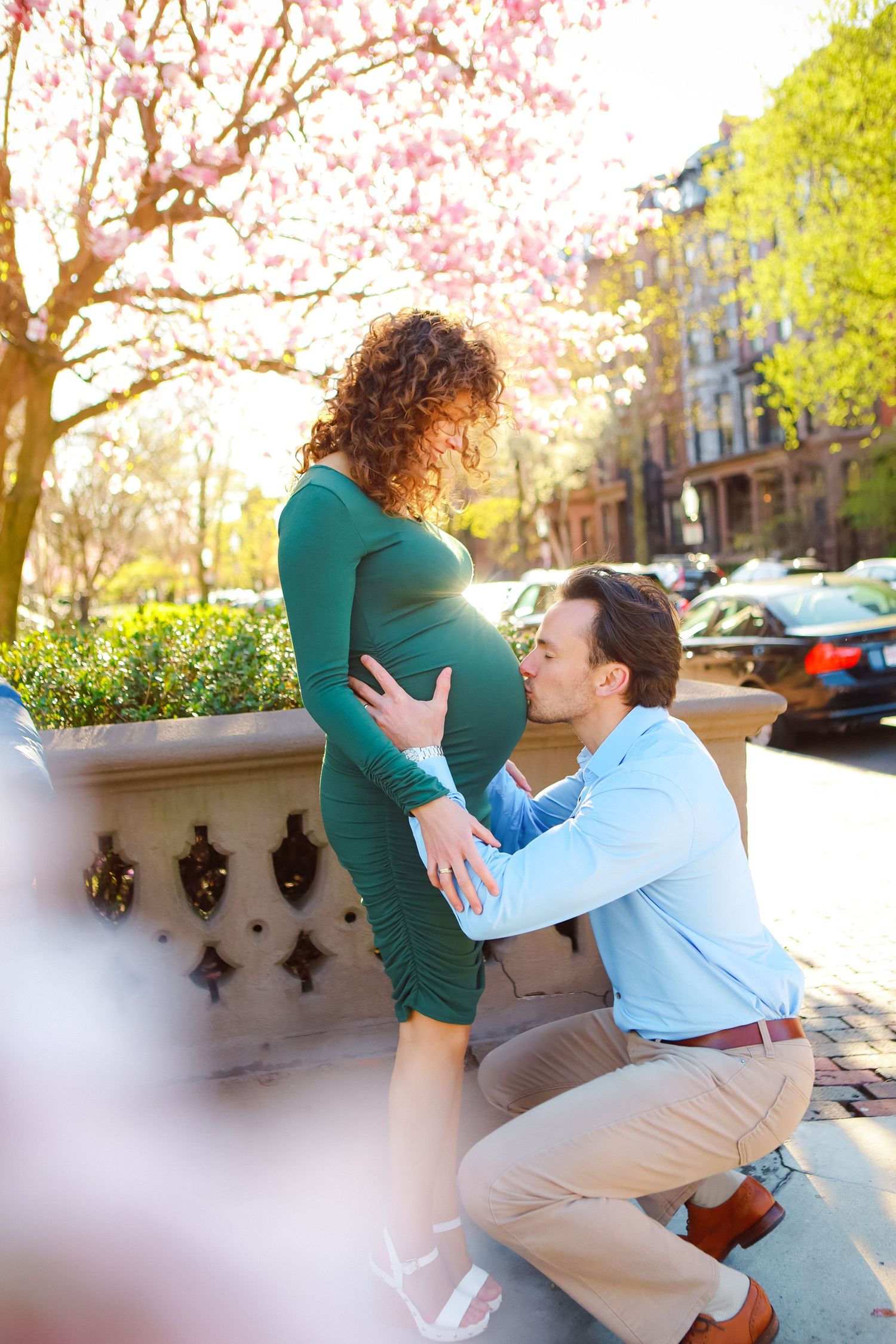 Man kneels to kiss pregnant womans belly near blooming magnolia tree on sunny urban street.