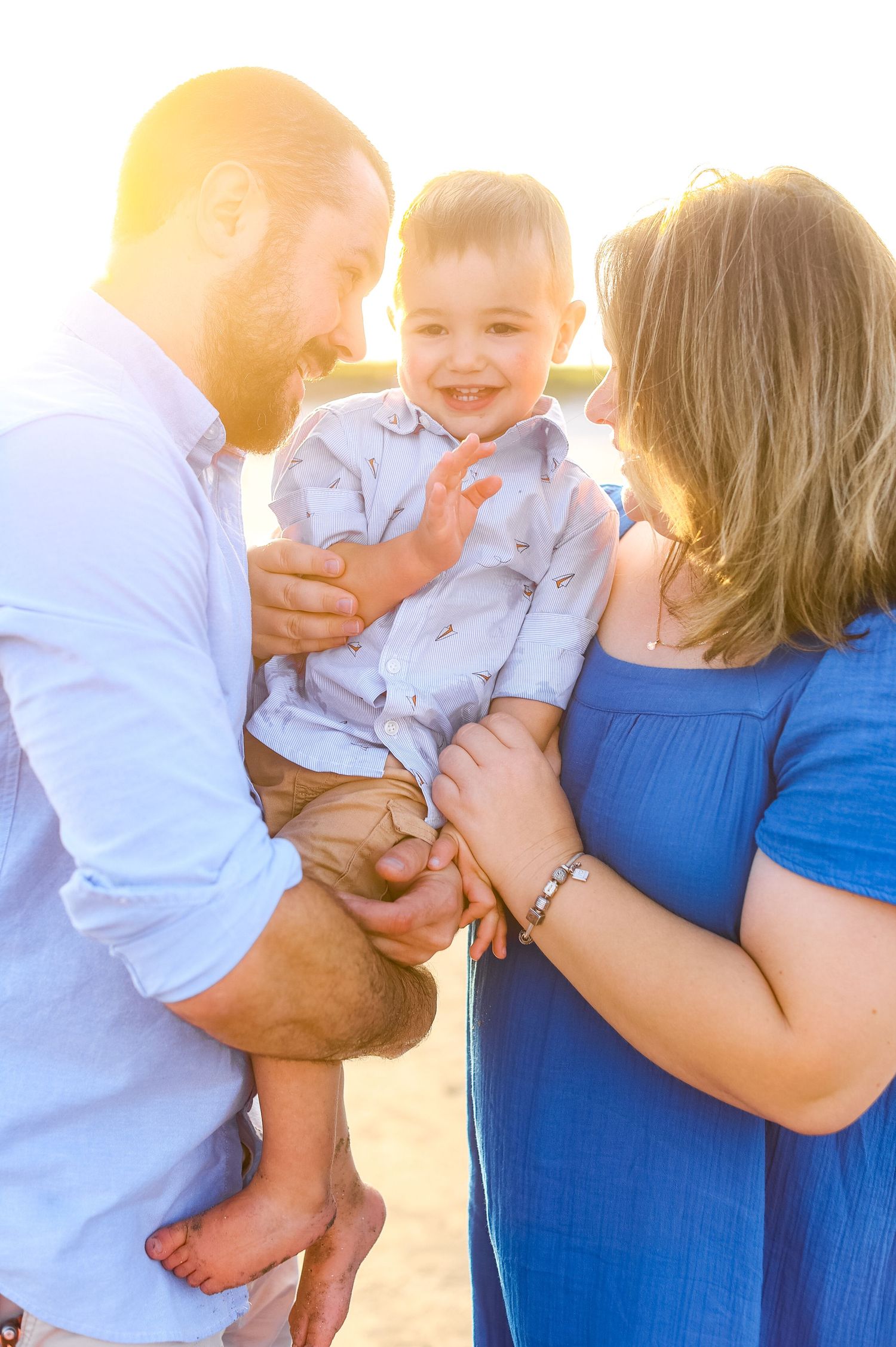 Happy baby boy laughing while being held by parents on a sunny beach at golden hour.