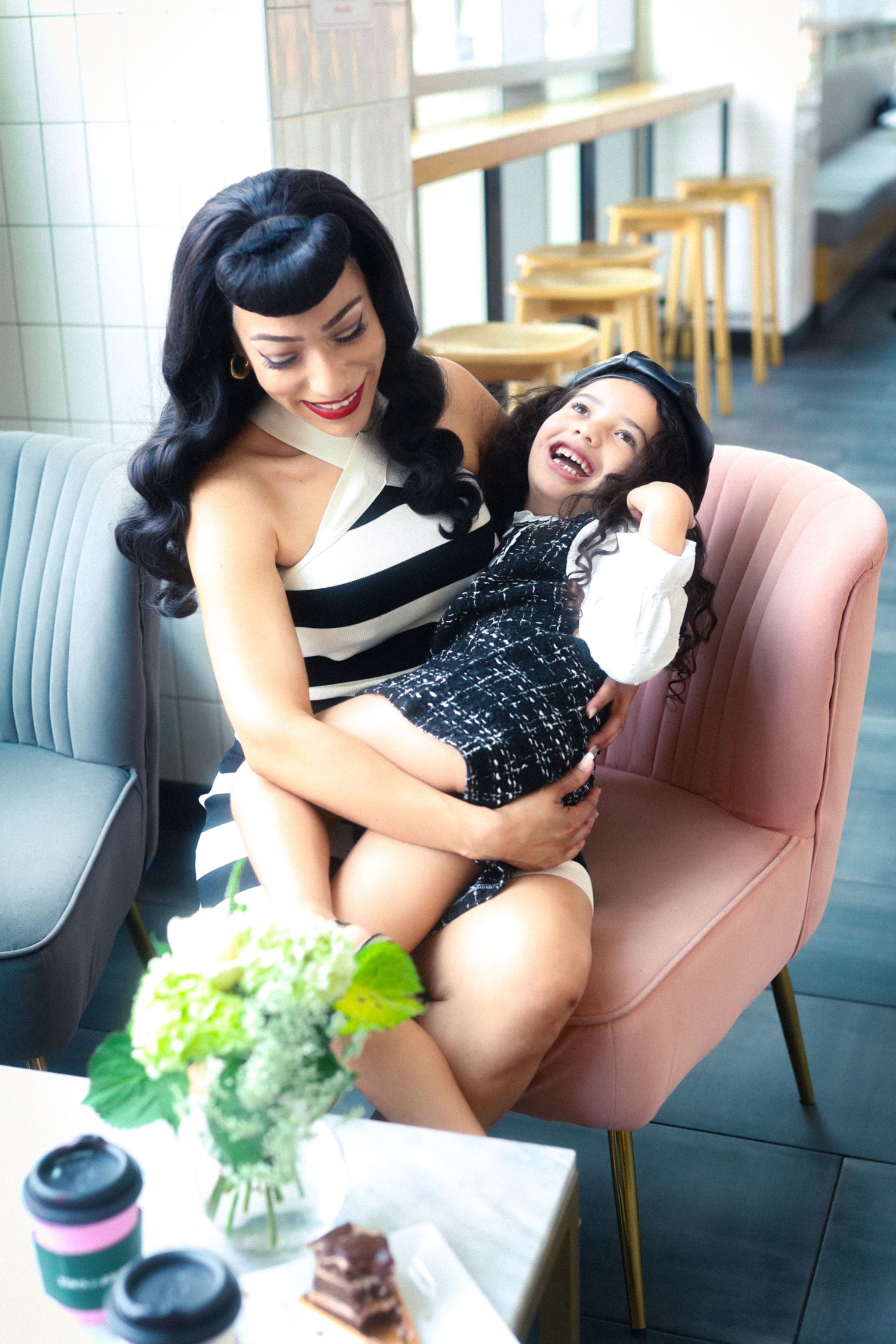 Stylish mother in striped dress laughing with young daughter on pink chair in cafe.