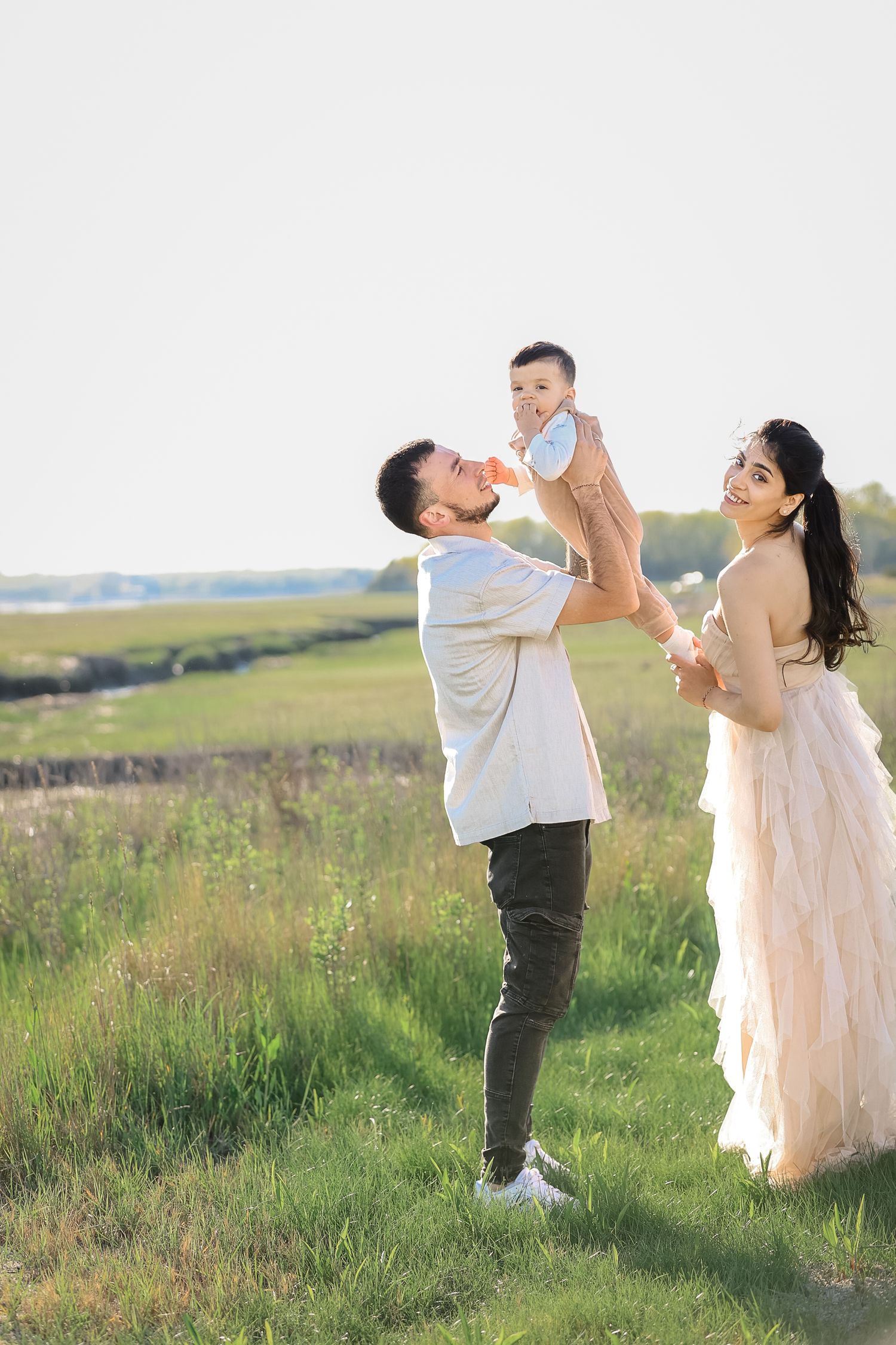 Father lifting baby while mother in pink gown smiles in field.