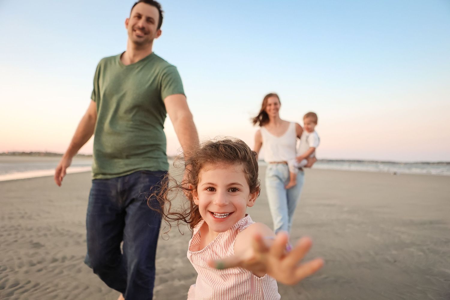 Joyful young girl reaching toward camera while family walks on beach at sunset.