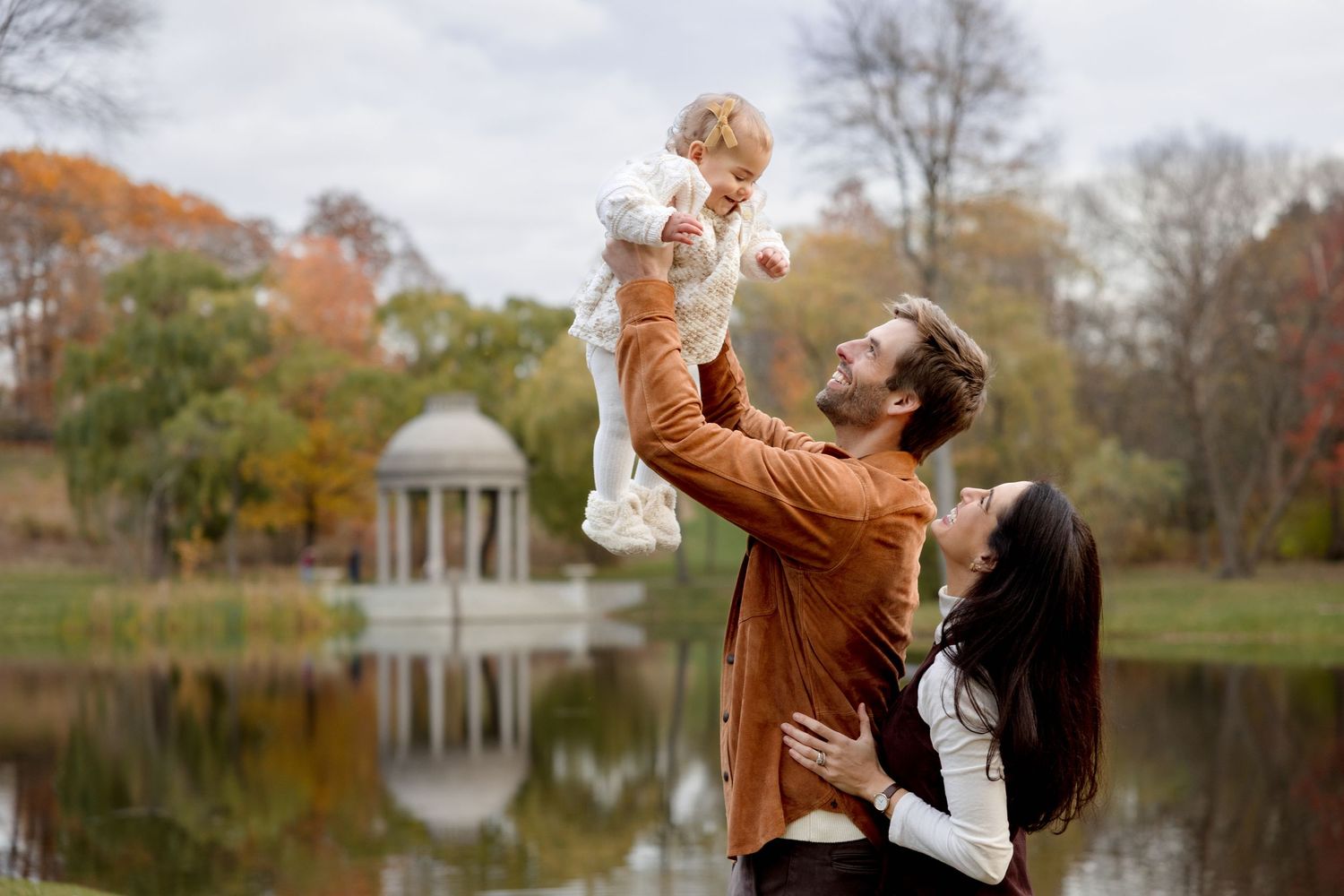 Young couple lifting baby in autumn park by lake with gazebo.