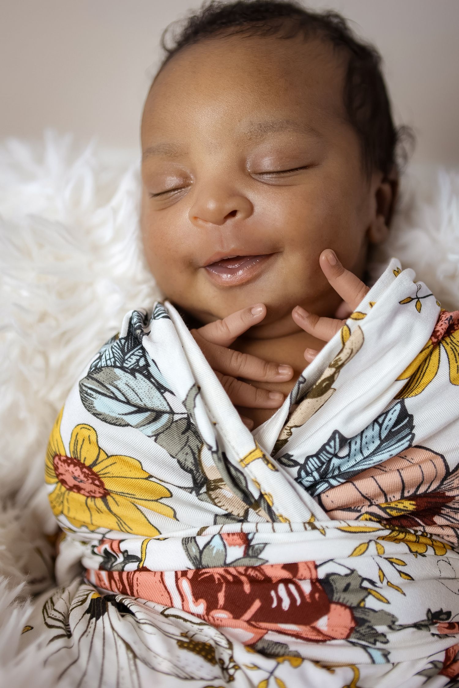 Smiling newborn baby swaddled in a colorful floral blanket on a white fluffy surface.