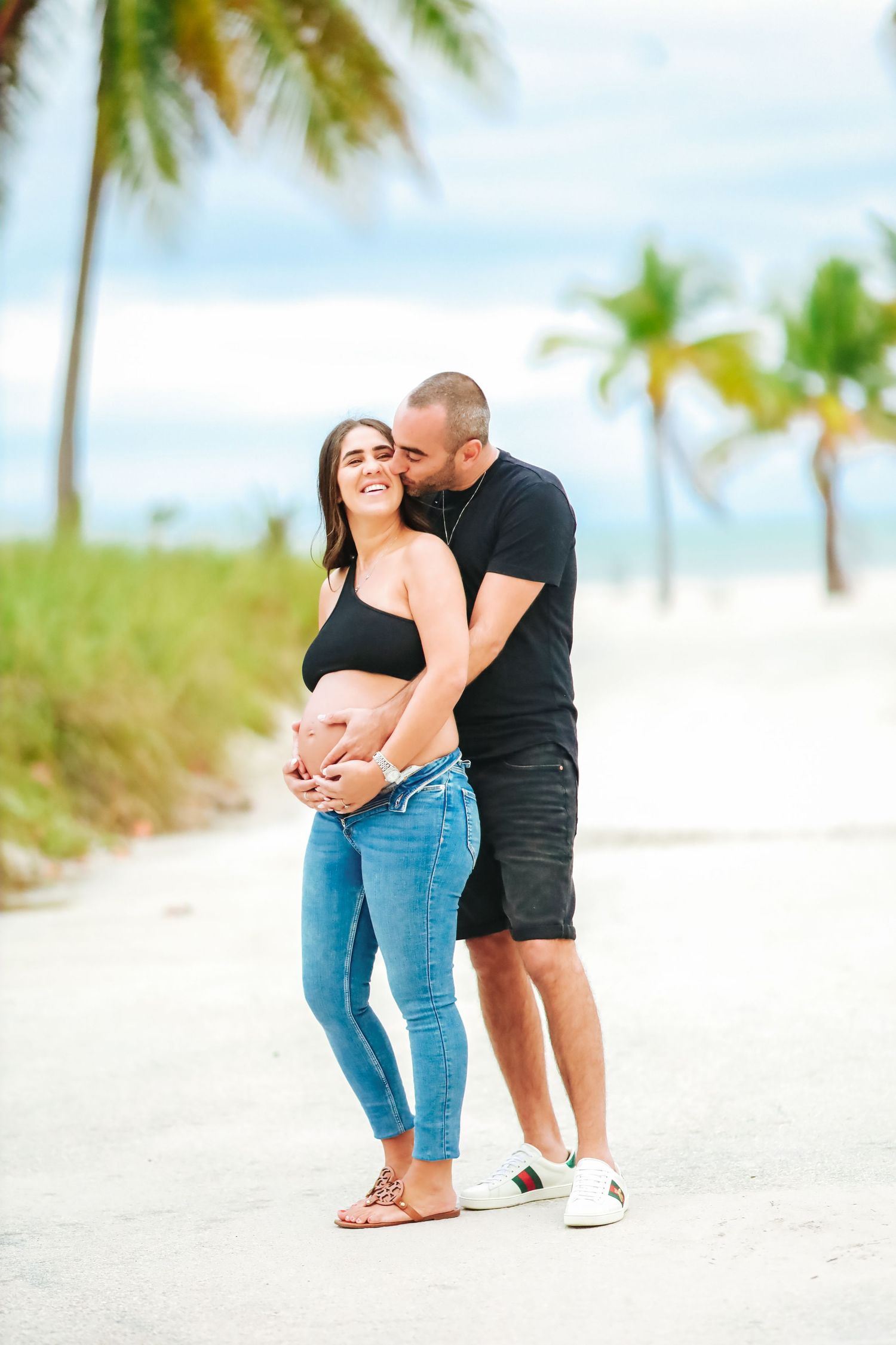 Man kissing pregnant woman on cheek while holding her belly on sunny tropical beach path.