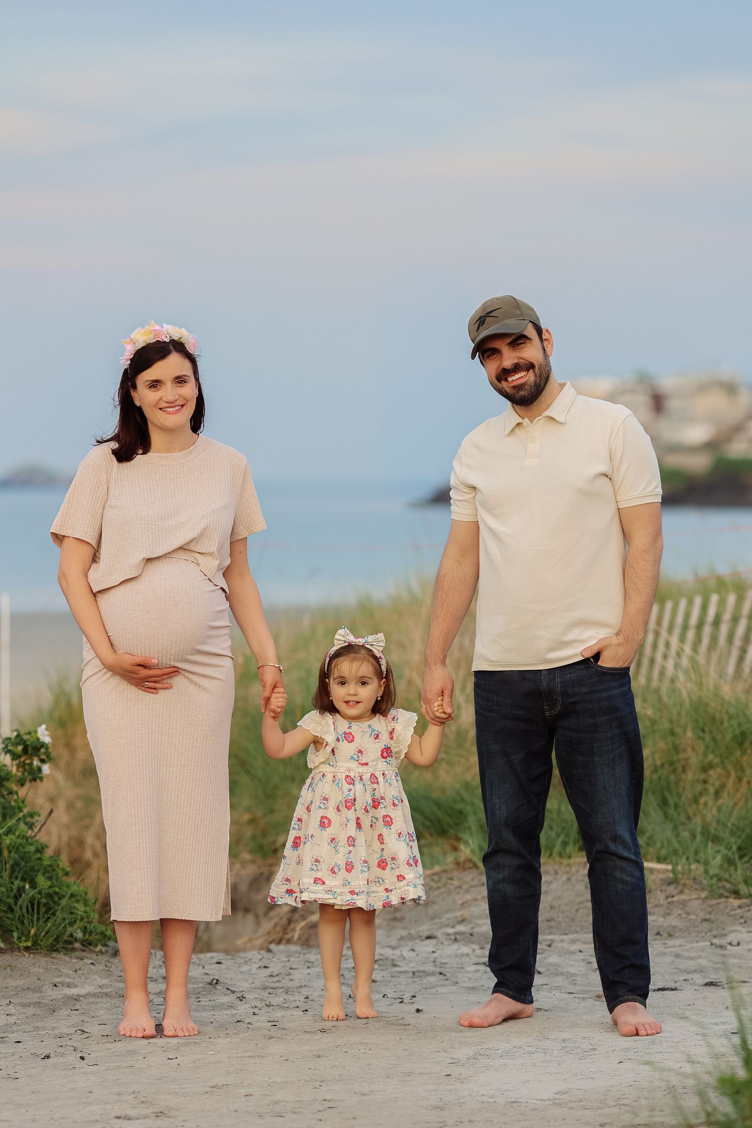 Pregnant woman, bearded man, and young daughter hold hands barefoot on a sandy beach path.